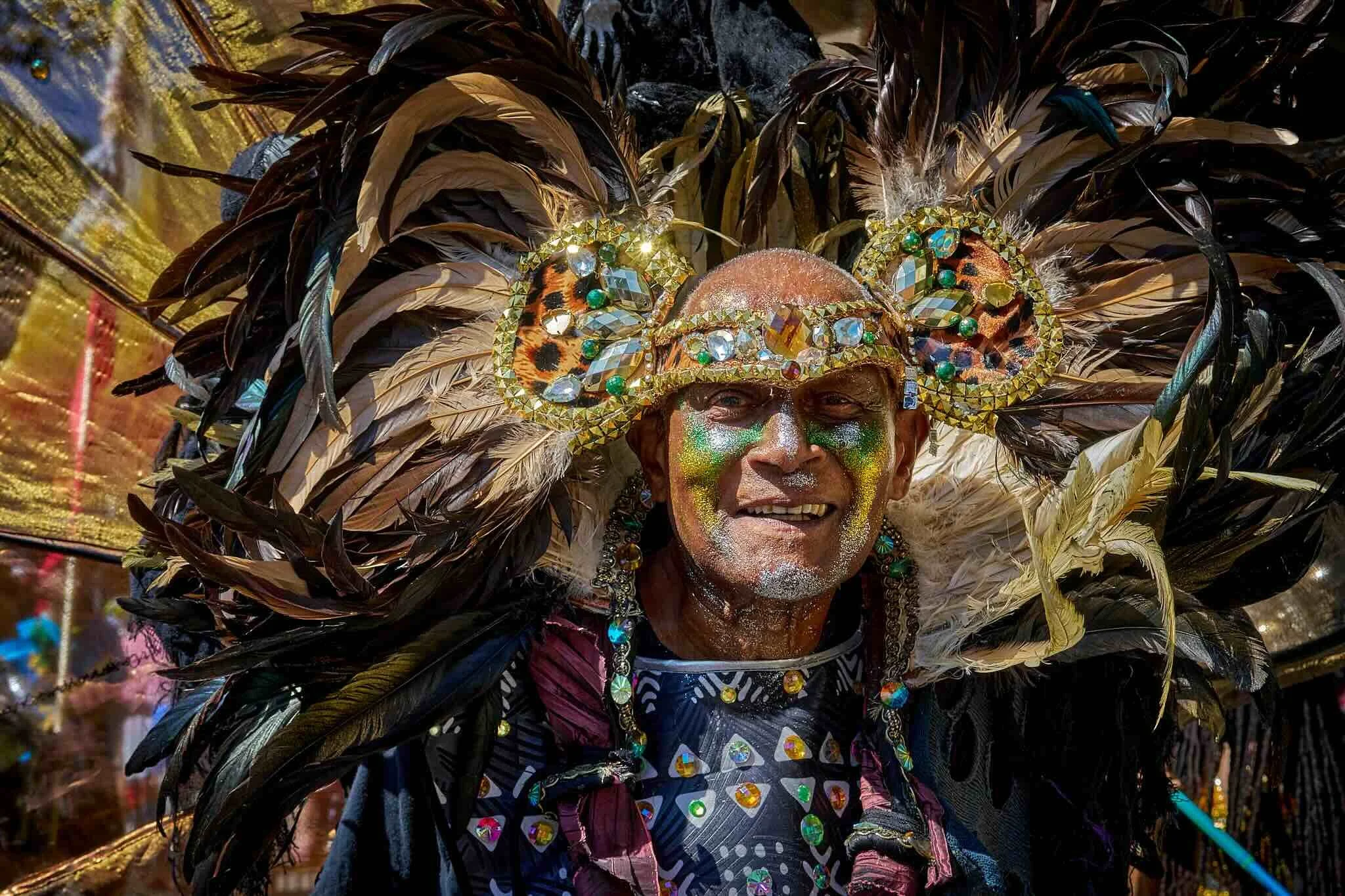 A person wearing an elaborate headdress made of large, dark feathers, decorated with shiny beads and jewelry, with face paint and glitter, smiling at a cultural celebration or festival.