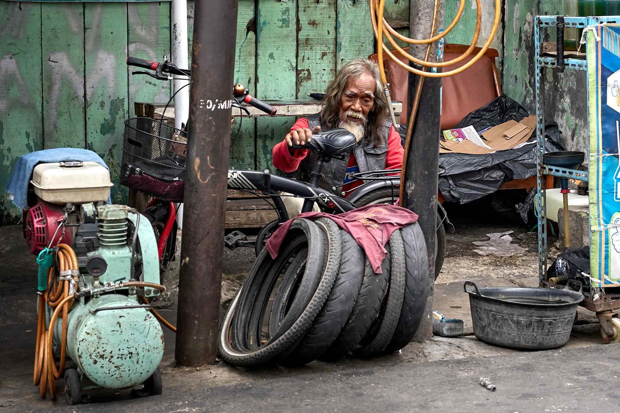 An elderly man with long gray hair and beard, wearing a vest and red shirt, sitting behind a bicycle in a cluttered outdoor space with tires, a small engine, and various tools and supplies in front of a weathered green wooden wall.