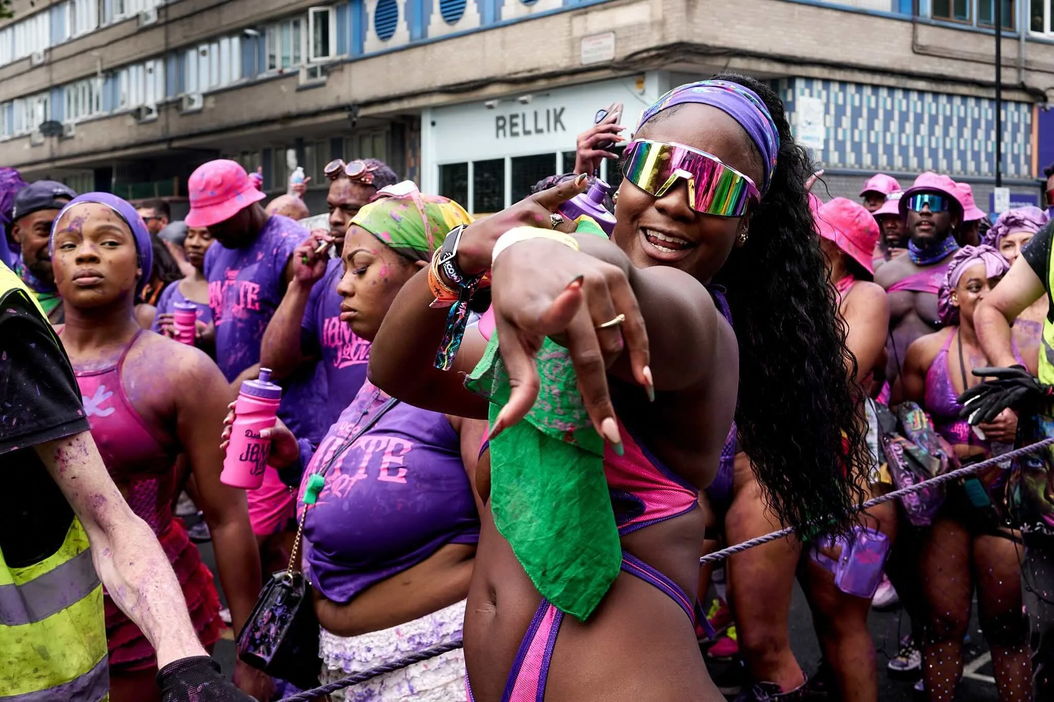 A diverse group of people celebrating in a colorful parade, wearing purple and pink outfits, some with paint on their skin, and holding spray bottles, in an outdoor urban setting.
