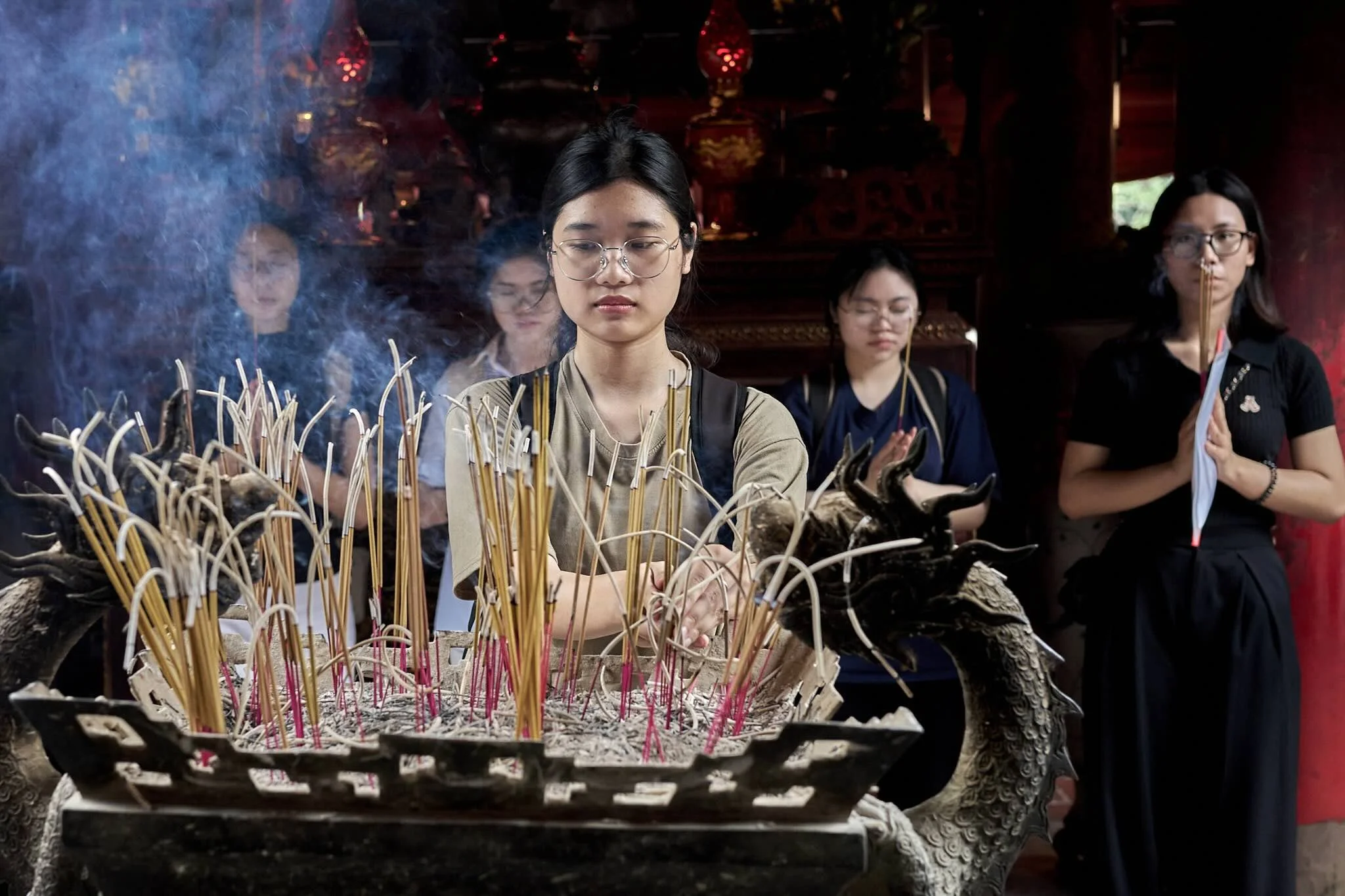 A group of women participating in a traditional Buddhist incense offering ceremony, with one woman in the foreground with closed eyes, surrounded by lit incense sticks, inside a temple with dark wooden interior.