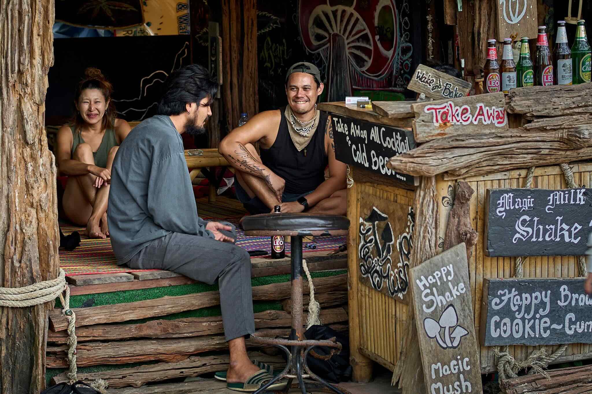 Three people sitting inside a rustic beer stand, two men and a woman, smiling and talking, with a background of colorful signs and bottles, wooden accents, and outdoor setting.