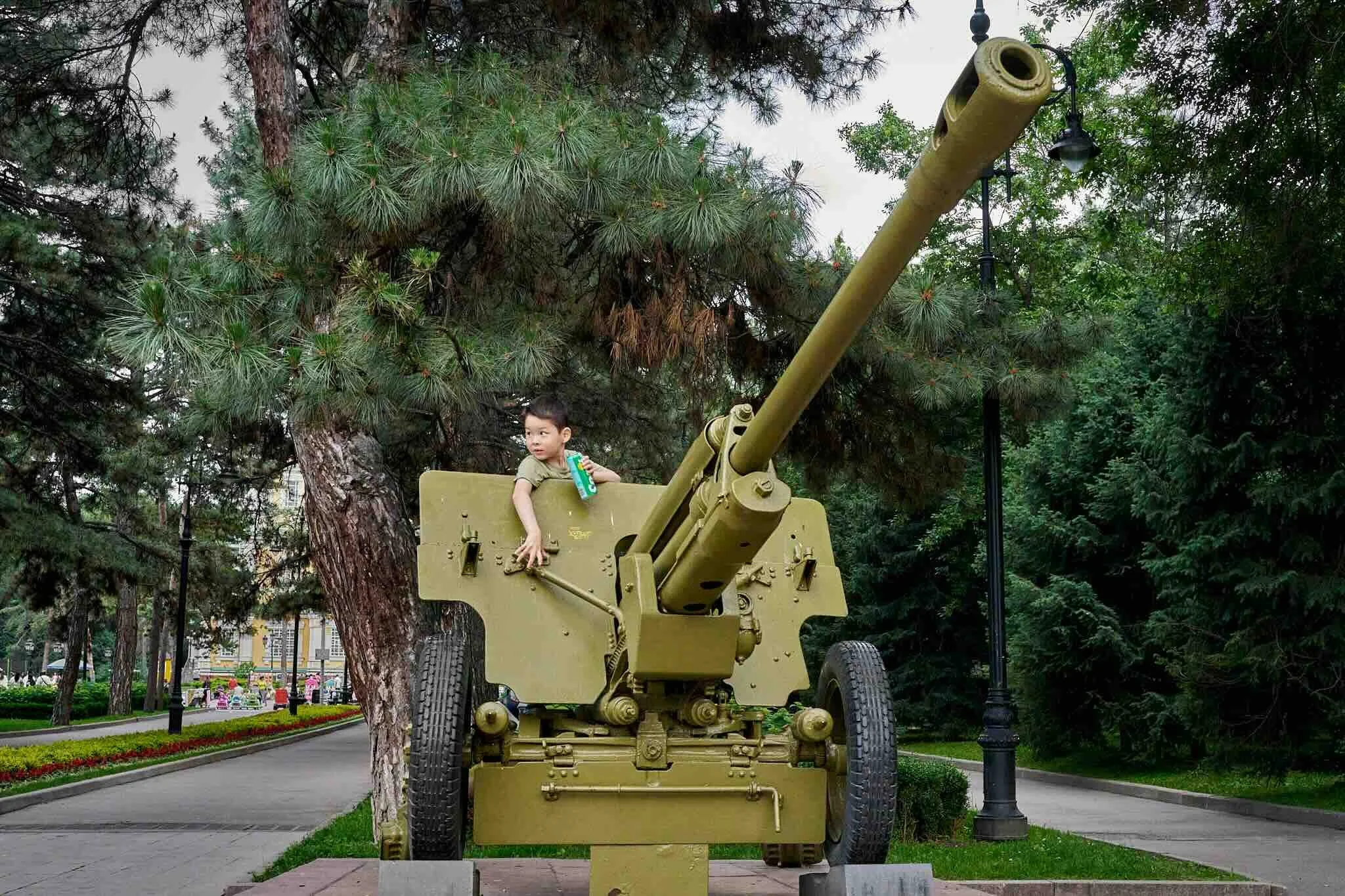 Jerzy Jaworek, Street and Travel Photography, Kazakhstan, Almaty. A young boy sitting in the turret of a large, yellow military tank in a park with trees and lampposts in the background.
