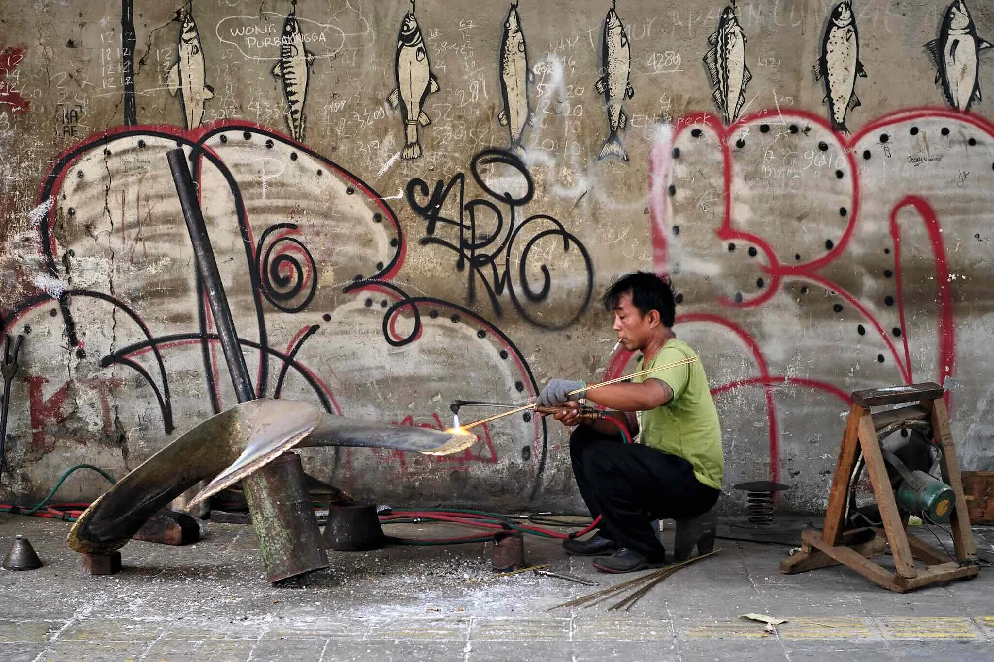 A man appears to be working on metalwork in an industrial setting with a concrete wall decorated with drawings of fish and chalk marks behind him. He is kneeling on the ground, holding a rod with tongs, and sparks are flying from the metal piece he i
