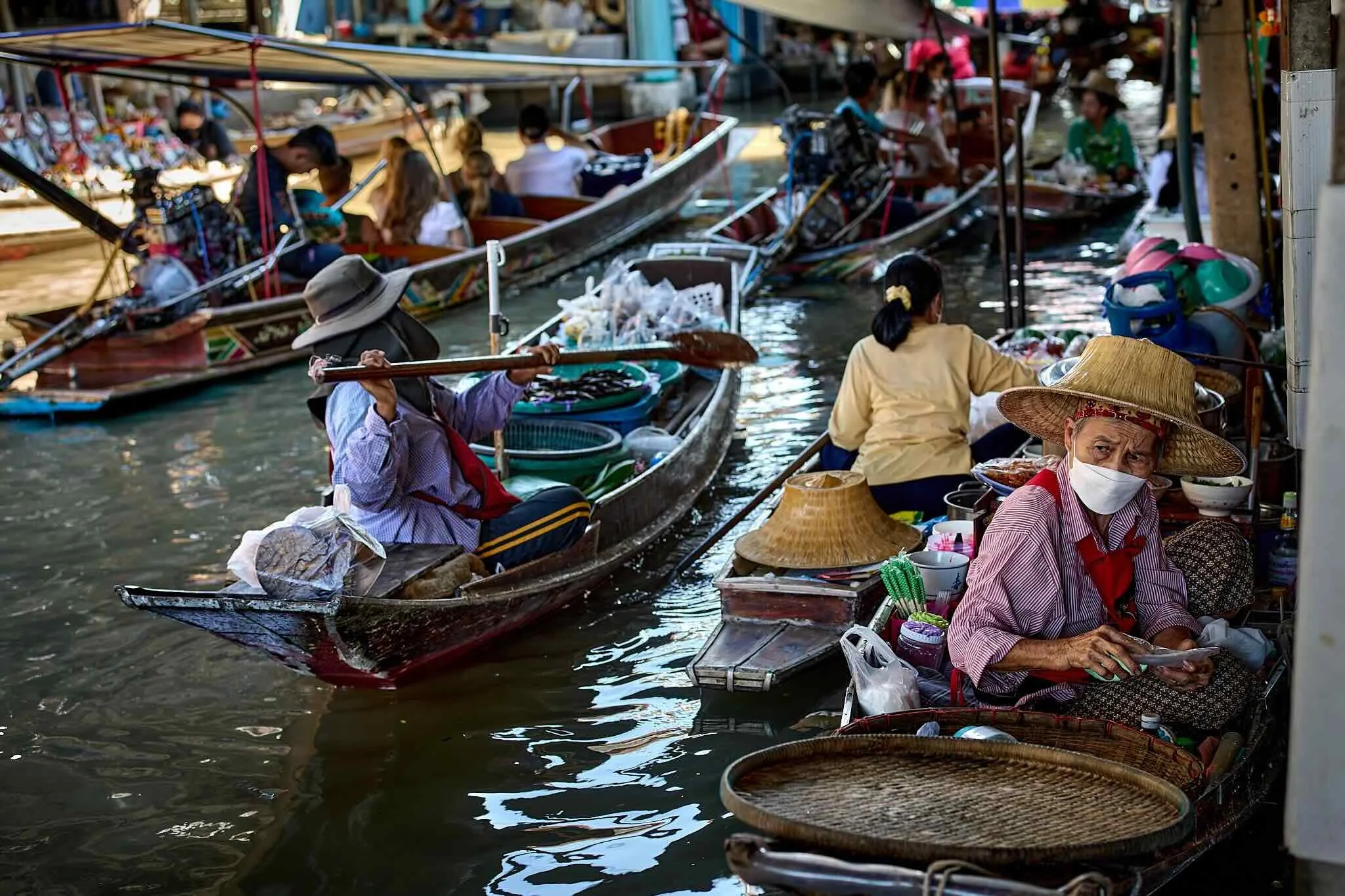 People in boats at a floating marketplace, selling goods along a waterway with vendors wearing hats and masks.