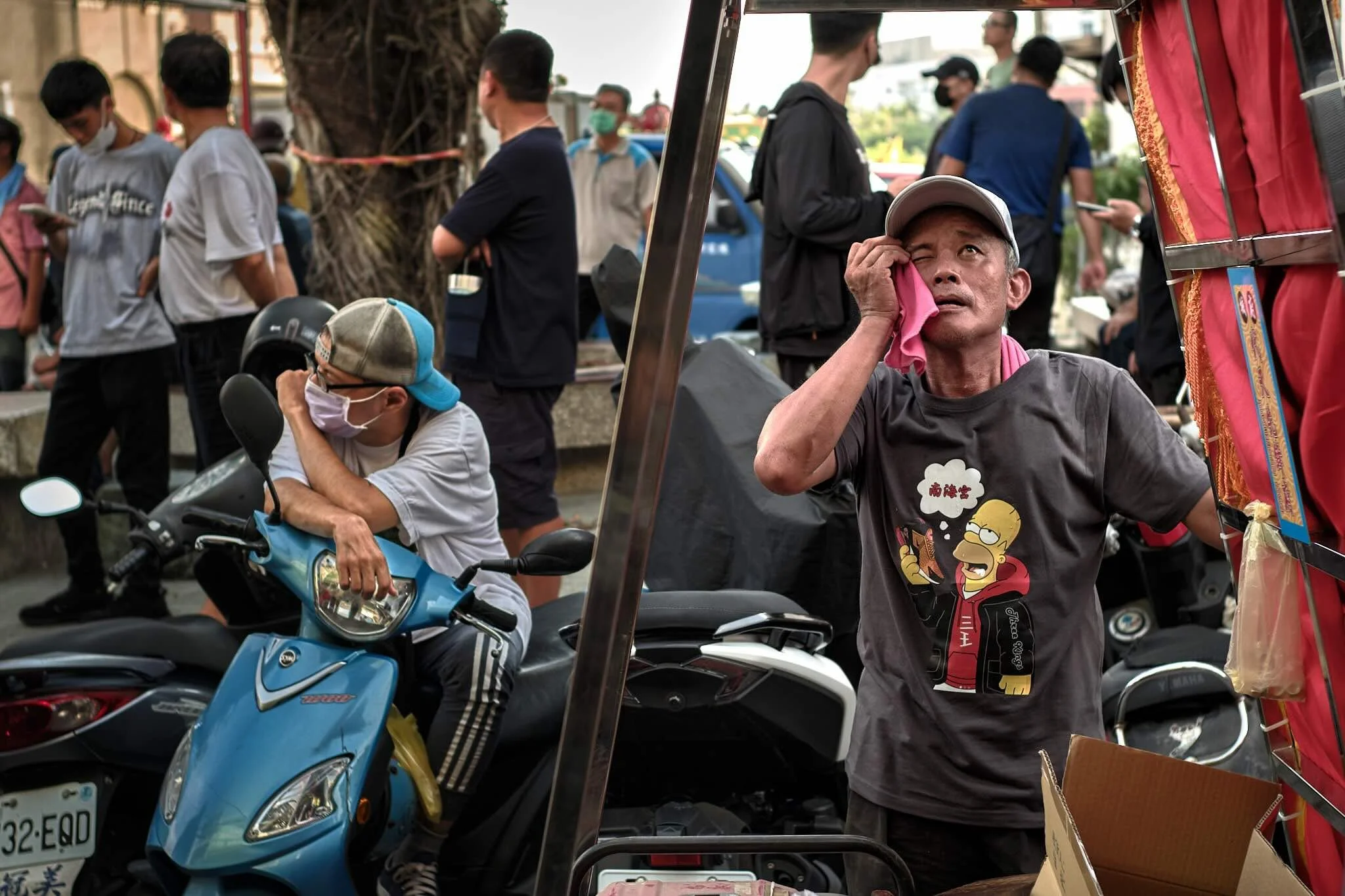 A man in a black 'Homer Simpson' t-shirt, hat, holding a pink cloth to his eye, sitting next to a motor scooter, with a busy outdoor setting of people and street vendors in the background.
