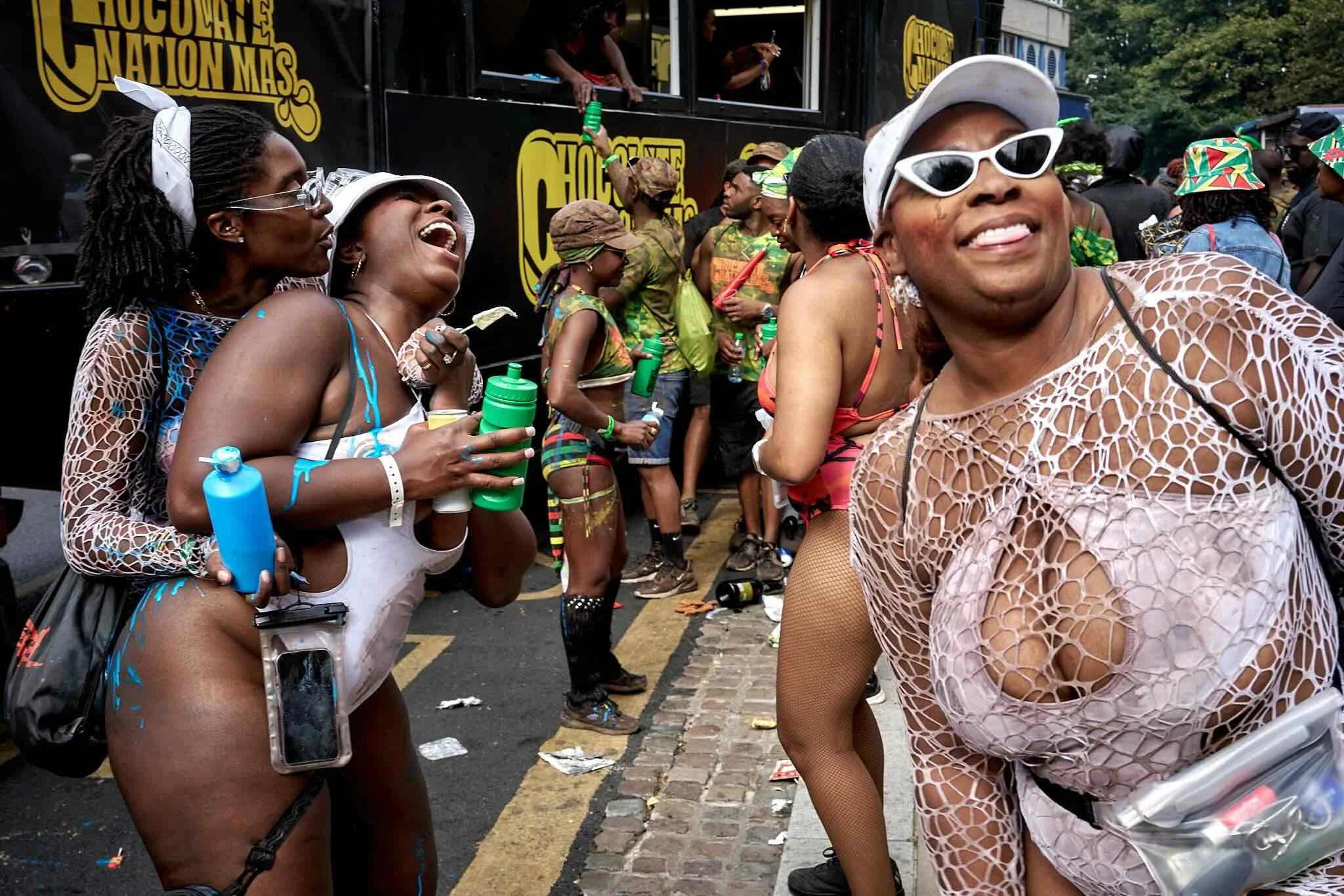 People celebrating at an outdoor event, some wearing colorful costumes and accessories, with a truck in the background featuring a yellow logo that reads 'Chocolate Nation MAS.' They are smiling, laughing, and enjoying the festivities.