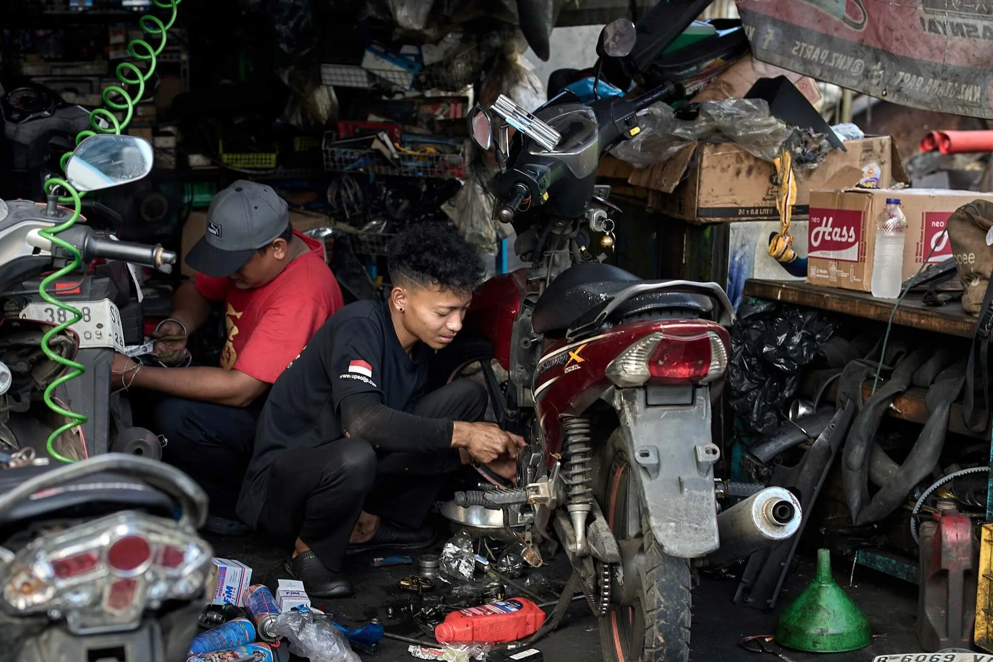 Two men working on a motorcycle in a cluttered workshop with shelves and boxes around them.
