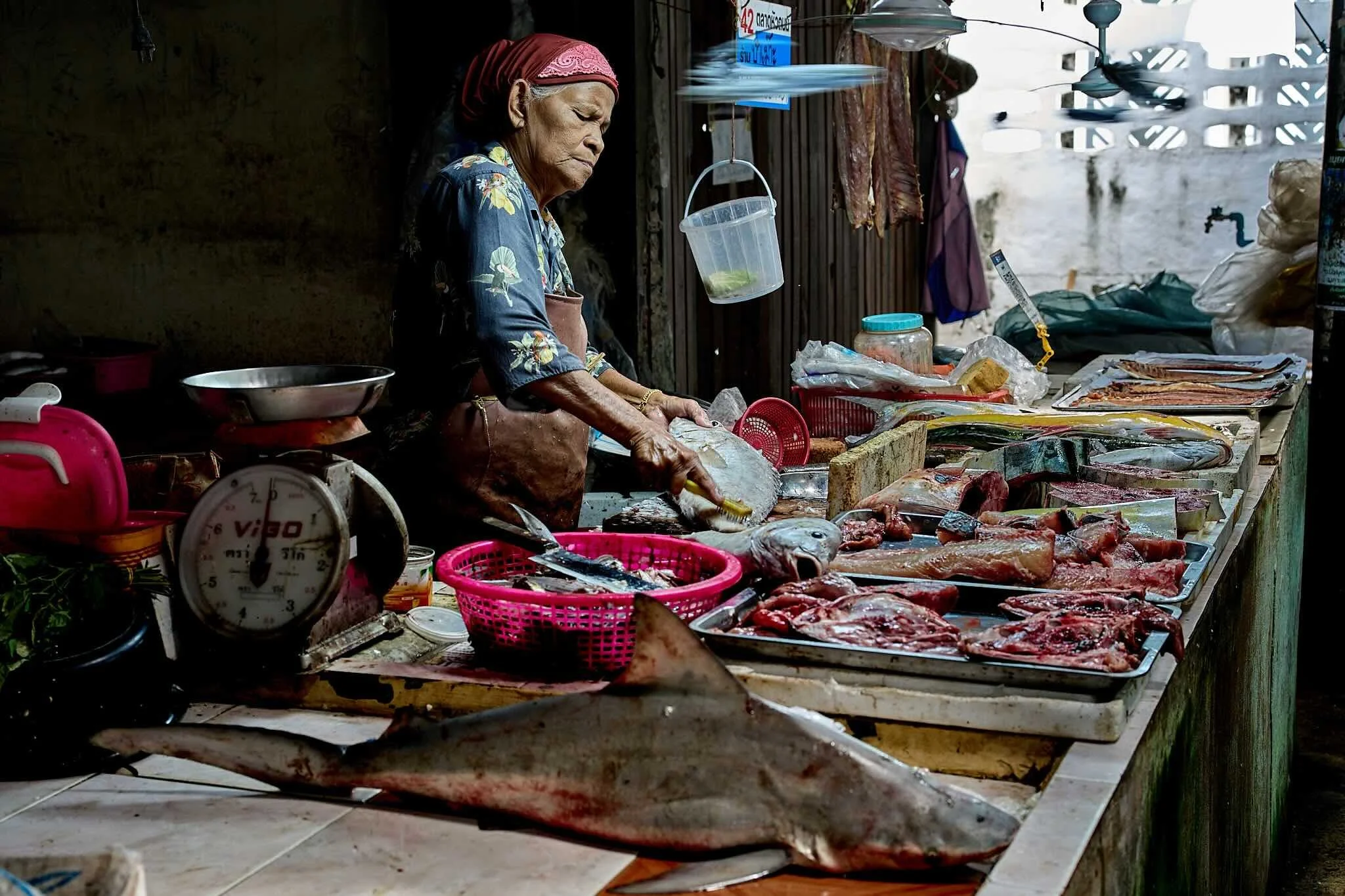An elderly woman wearing a red headscarf and floral shirt preparing fish at a market stall with various cuts of fish and seafood displayed on the counter.