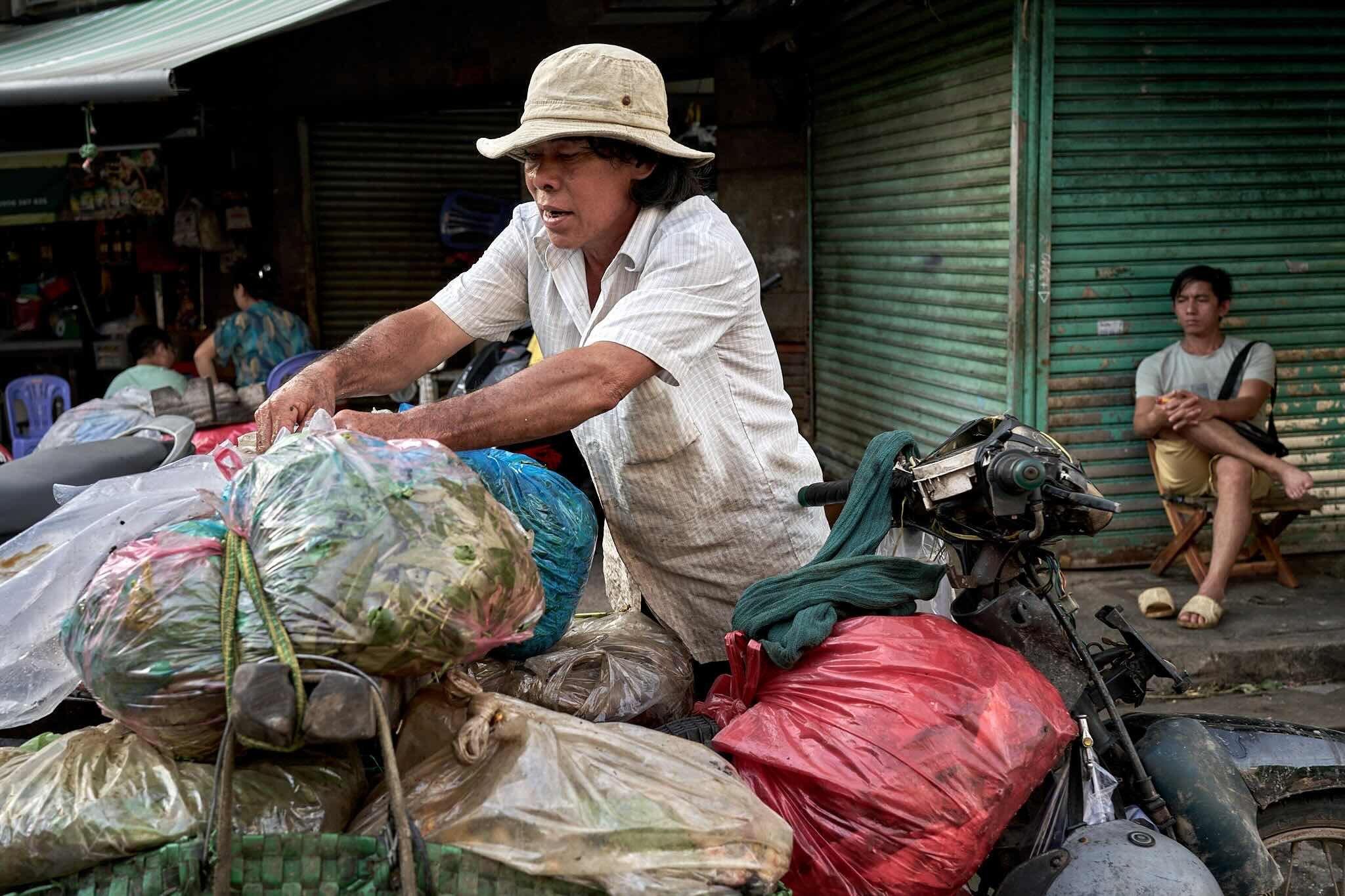 A man wearing a beige wide-brimmed hat and a white shirt is leaning over a pile of plastic bags filled with various items, standing next to a motorbike. In the background, there are people sitting and a closed green metal shutter.