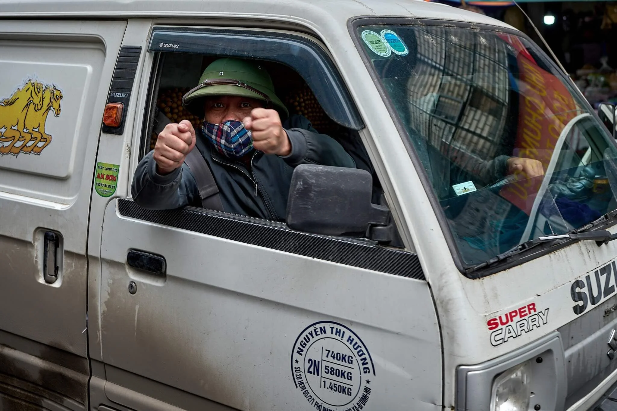 A man wearing a face mask and military-style hat sitting in the driver's seat of a white Suzuki truck decorated with various stickers and signs.