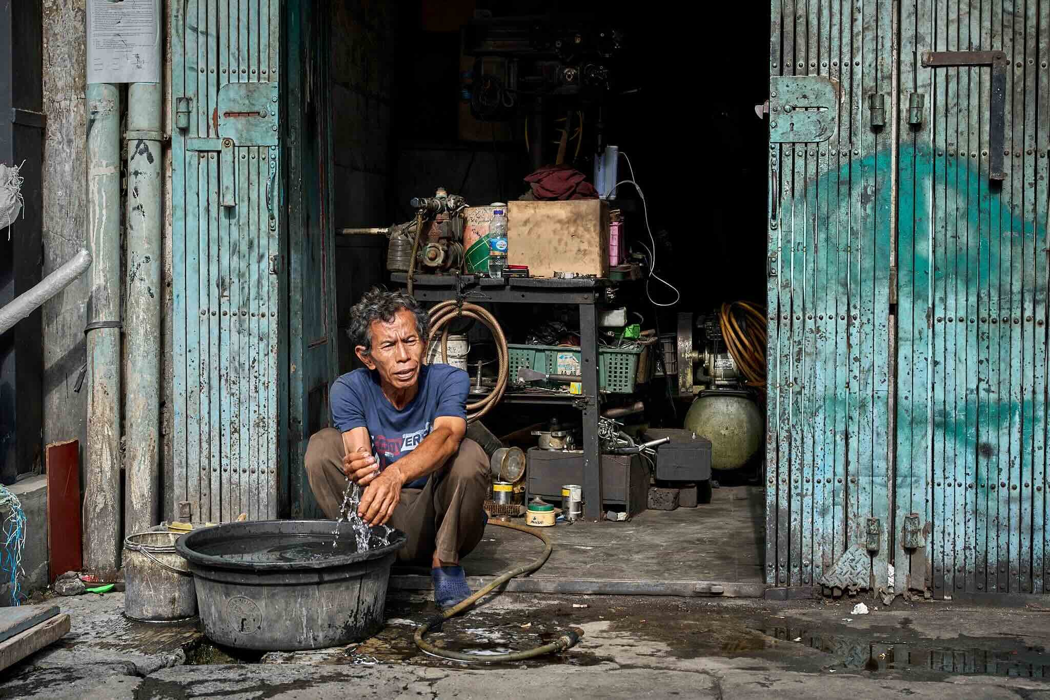 A man sitting outside in front of a rustic, teal-colored metal door, washing his hands in a large black bowl. The scene depicts a modest, possibly urban setting with various objects and tools, including a water pump, hose, bottles, and containers, ar