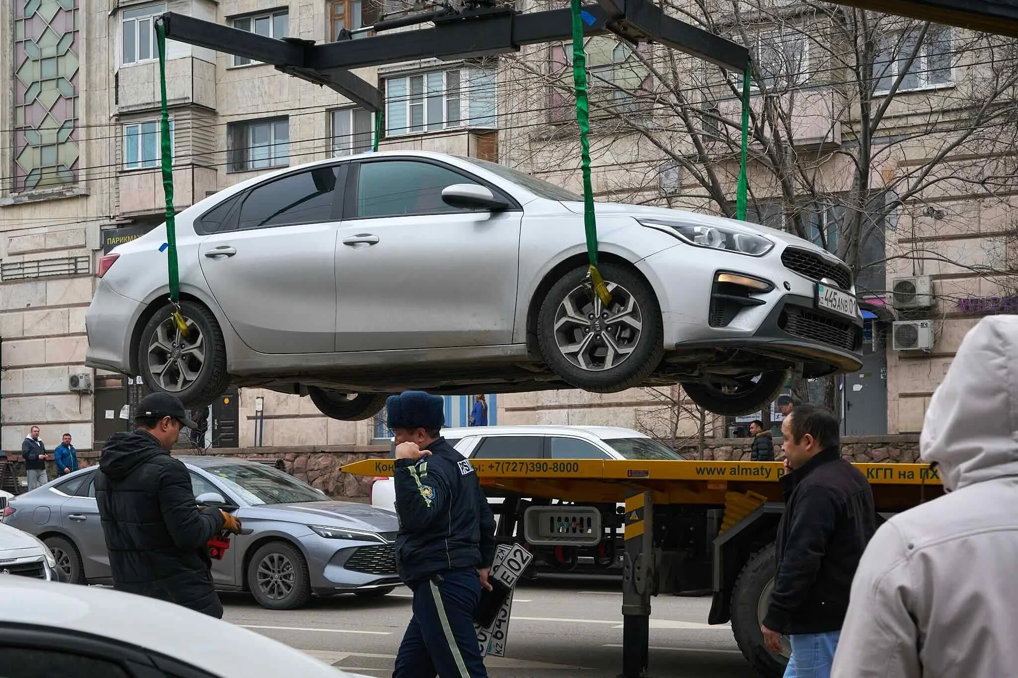 Jerzy Jaworek, Street and Travel Photography, Kazakhstan, Almaty. A white sedan car is lifted on a tow truck in an urban area with several people and onlookers around. Buildings and a tree are visible in the background.