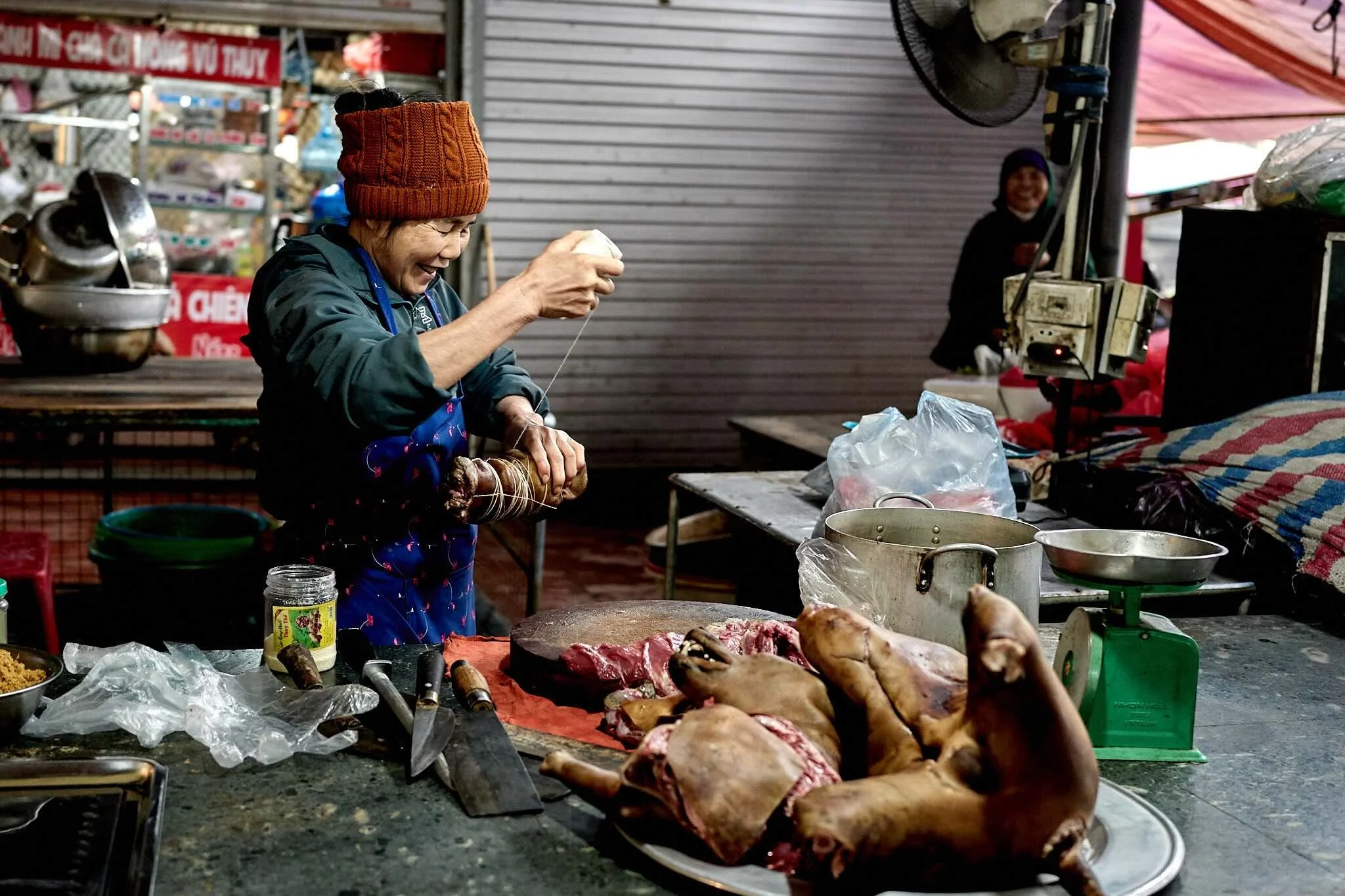 A woman smiling and preparing meat at an outdoor market stall with various tools and ingredients on the table.