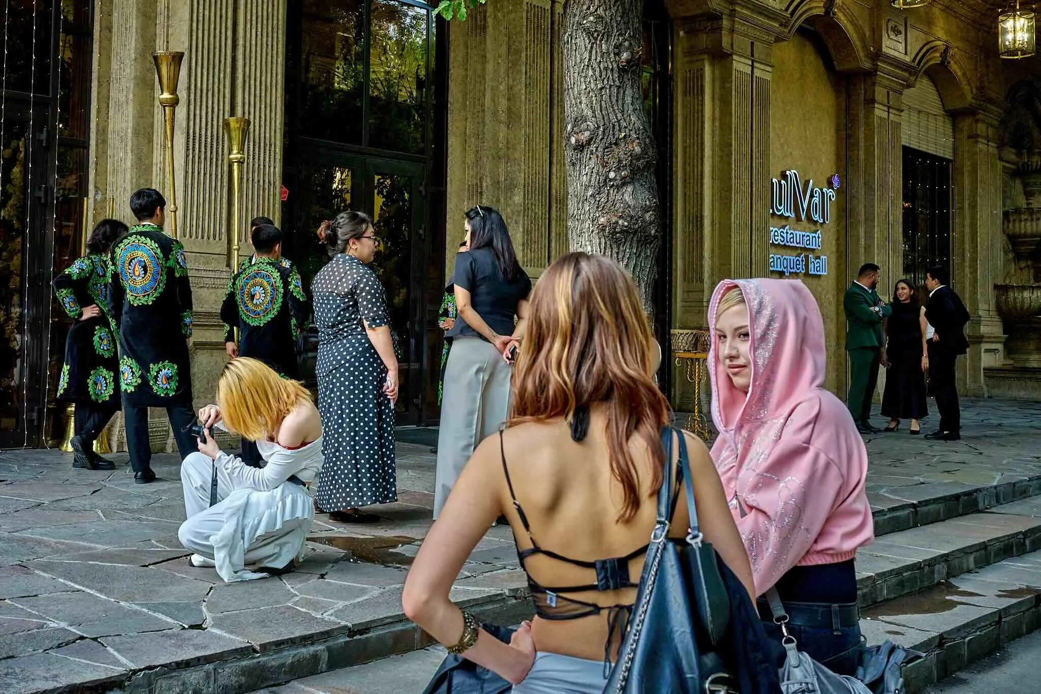 Jerzy Jaworek, Street and Travel Photography, Kazakhstan, Almaty.  People sitting and standing on a stone street outside a restaurant with a sign that reads 'Ulvar' and 'banquet hall', with a large tree and ornate building facade in the background.