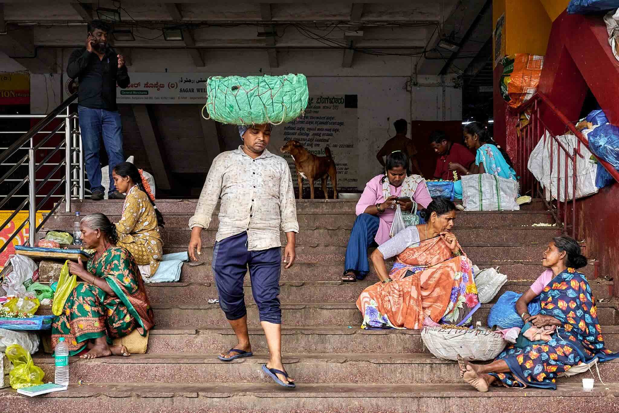 Jerzy Jaworek, Street and Travel Photography, India, Delhi. People sitting and standing on the stairs outside a building, some engaged in shopping or chatting, with a man walking down the stairs looking at his phone and several vendors selling goods.