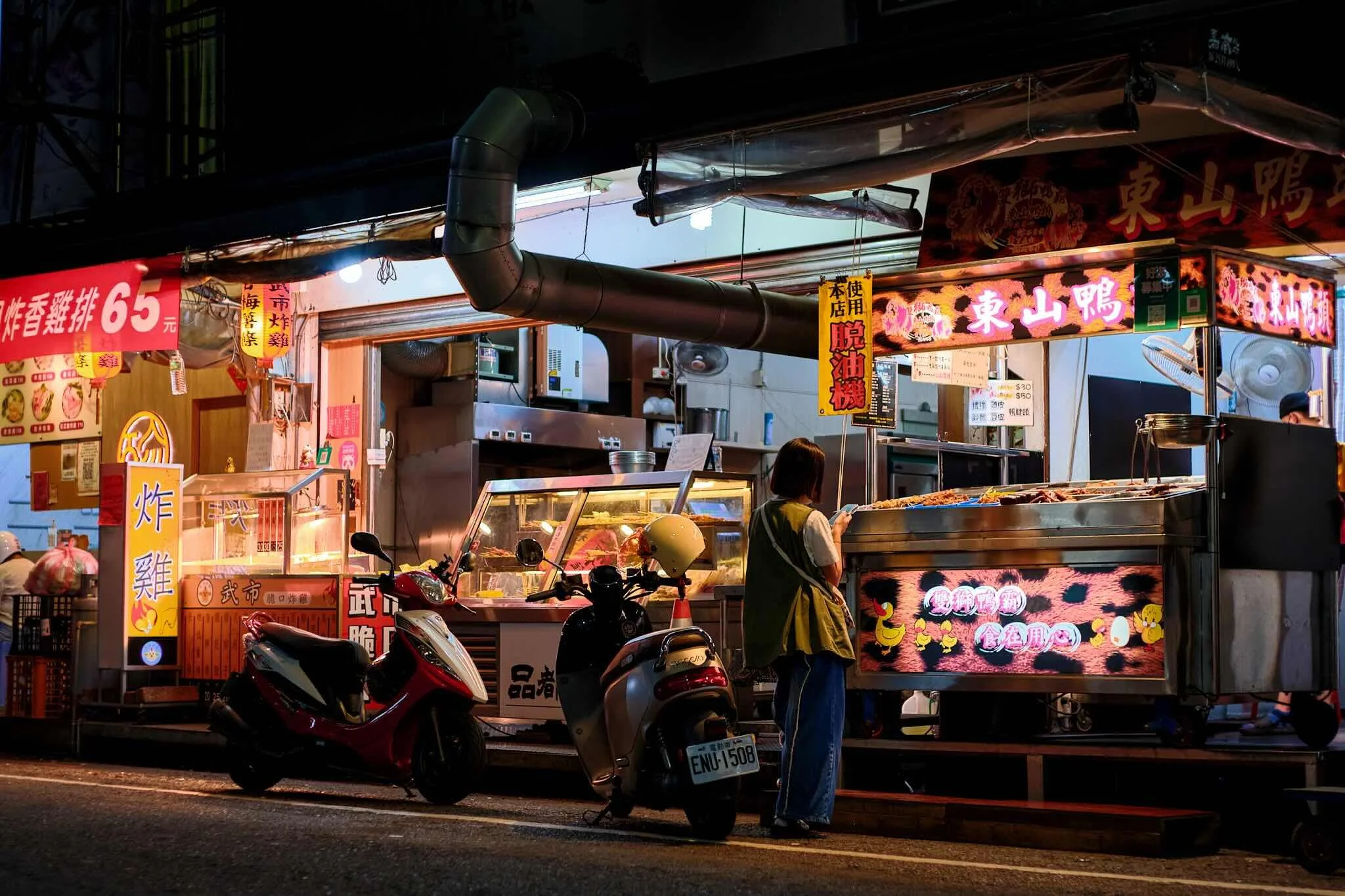 Night scene of a street food stall with illuminated signs, including Chinese characters, showcasing food items in a display case. Two motor scooters are parked in front, and a woman is standing nearby, possibly ordering or paying.