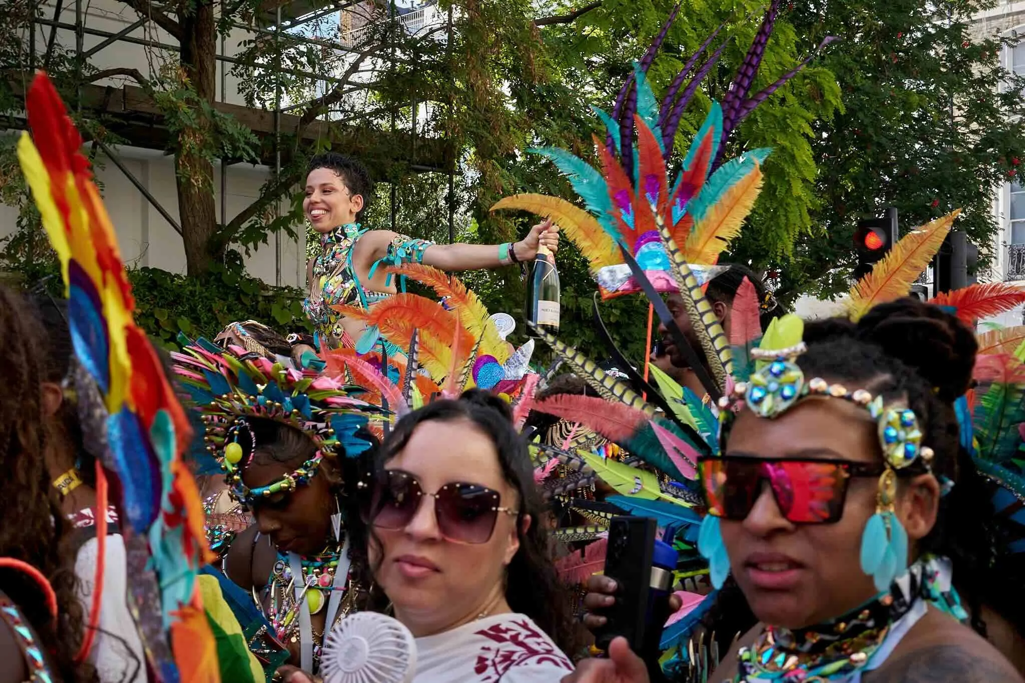 A group of people participating in a colorful parade or festival, wearing vibrant costumes with feathers and beaded accessories. One person is smiling and riding a float or parade vehicle, surrounded by others in elaborate outfits.