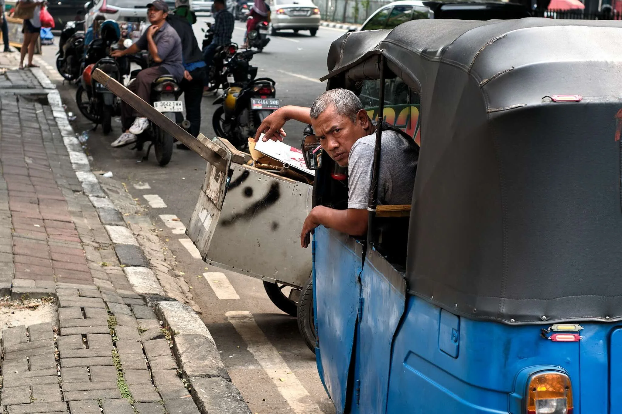 A man sitting in a small, covered blue and black tricycle vehicle, leaning his head and arm on the side, looking towards the camera. The vehicle is parked on a city street with motorcycles, cars, and pedestrians in the background.