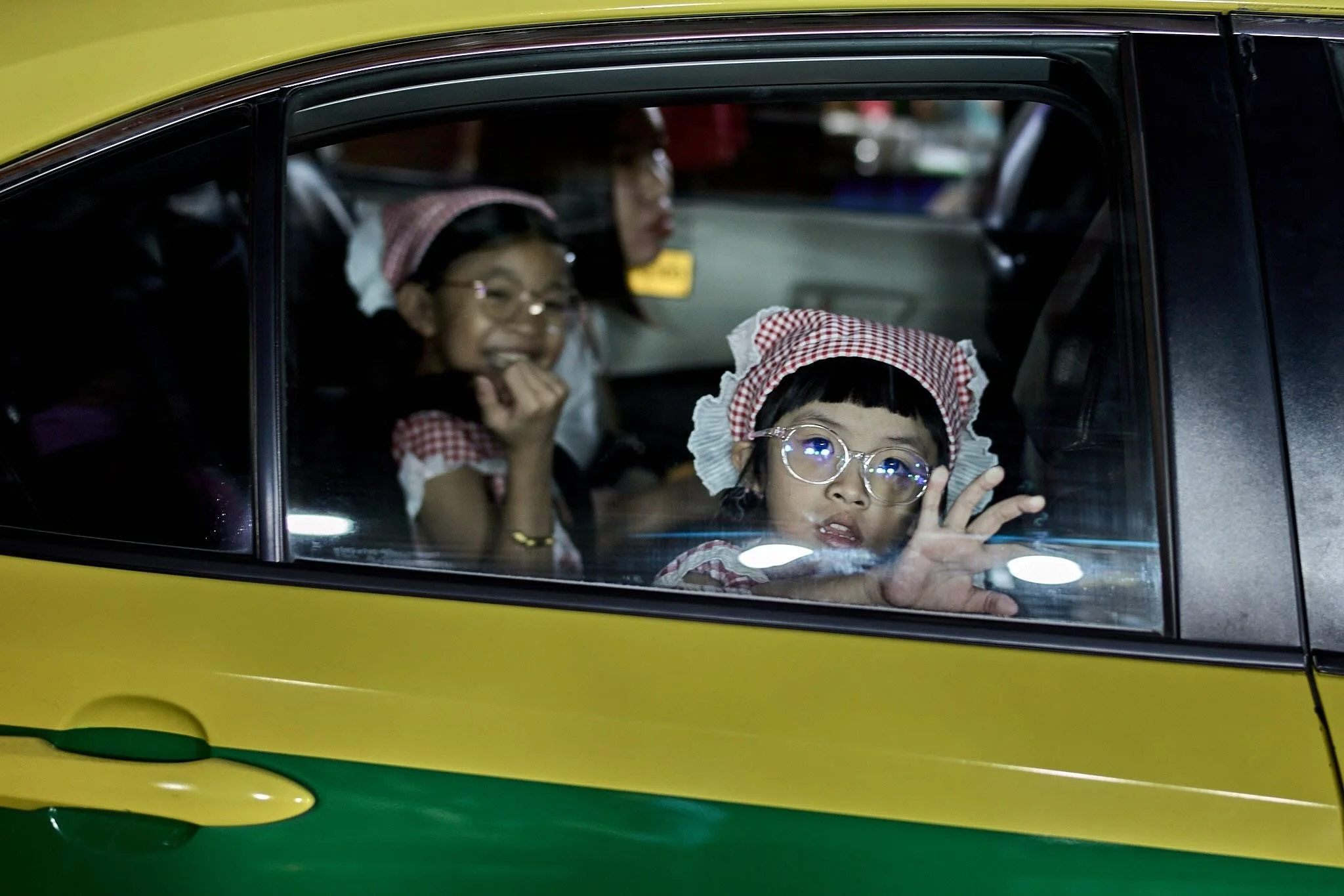 Two young girls wearing matching red and white checkered outfits and bonnets, smiling and waving from inside a yellow and green vehicle.