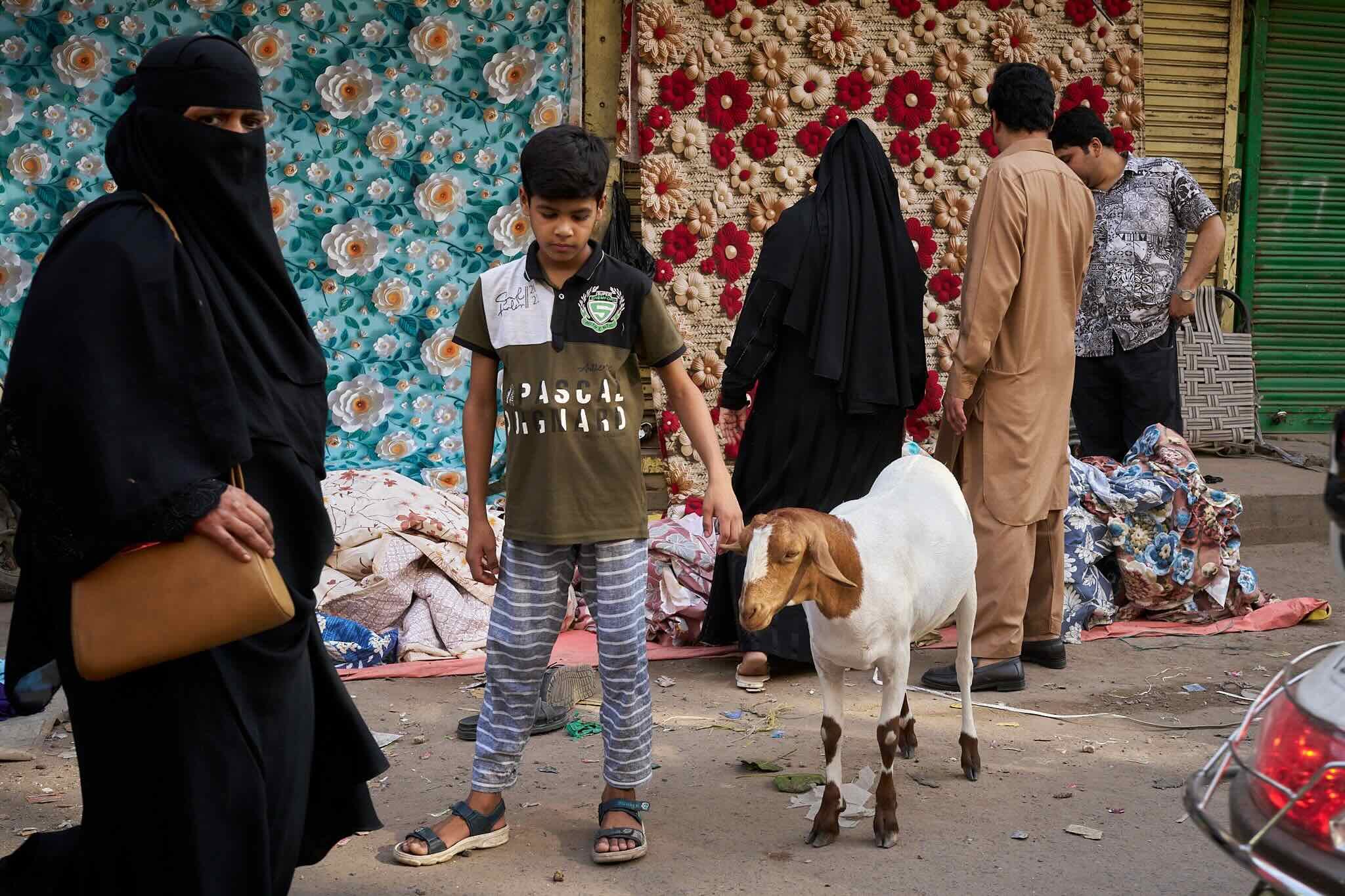 Jerzy Jaworek, Street and Travel Photography, India, Delhi. People gathered in an outdoor market with floral decorations in the background. Women and children, including a boy, are present, wearing casual and traditional clothing.