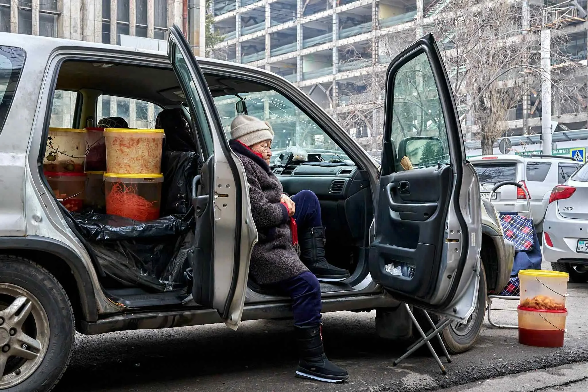Jerzy Jaworek, Street and Travel Photography, Kazakhstan, Almaty. An elderly woman sits inside a small, silver vehicle with the driver's side door open. The vehicle appears to be in a market or street setting with various containers, a small table, a