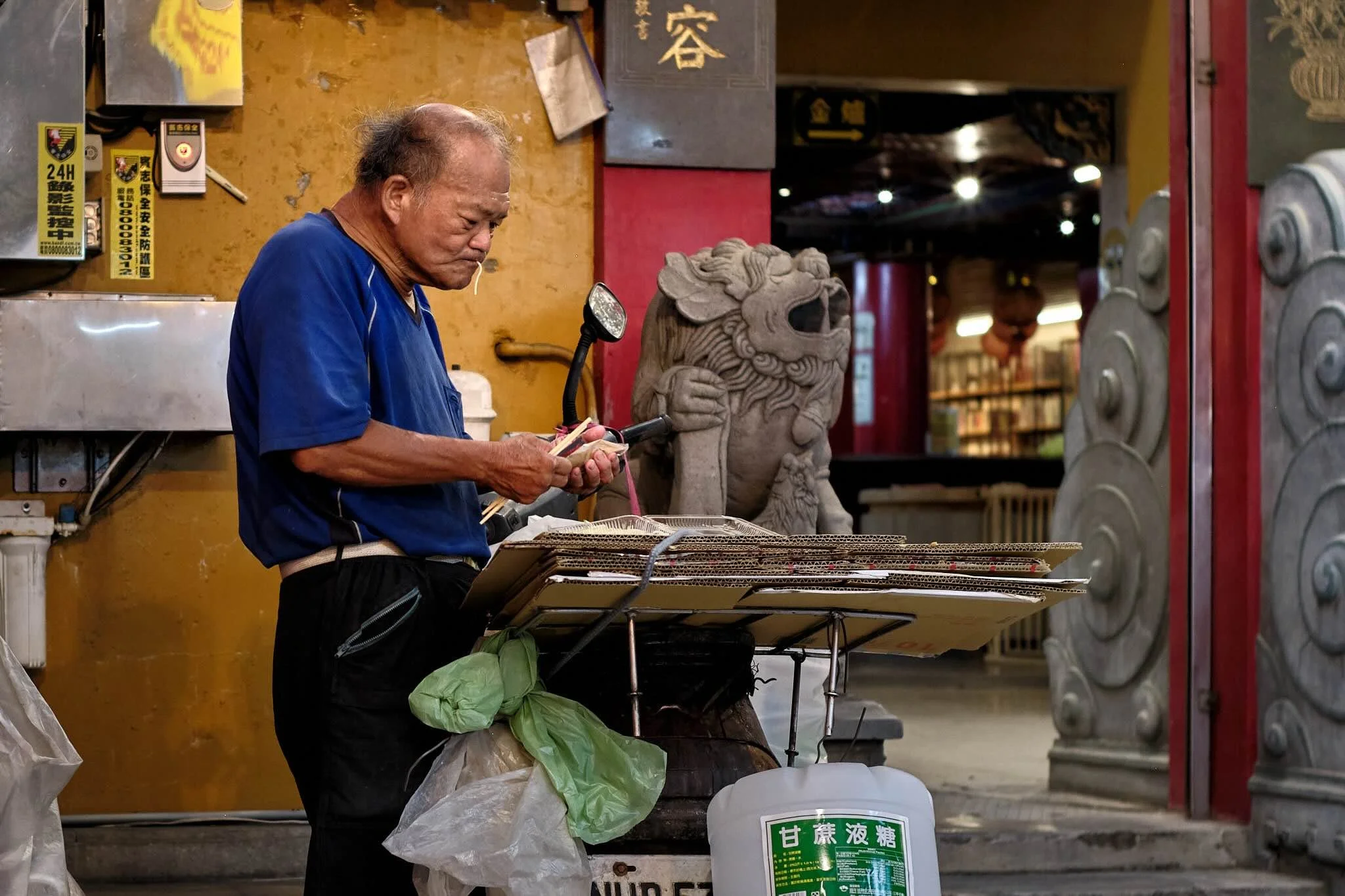 An elderly man with thinning hair and a serious expression stands next to a stone sculpture of a Chinese guardian lion inside a store. He is looking down at his hands, holding chopsticks, with various posters and signs on a yellow wall behind him, an