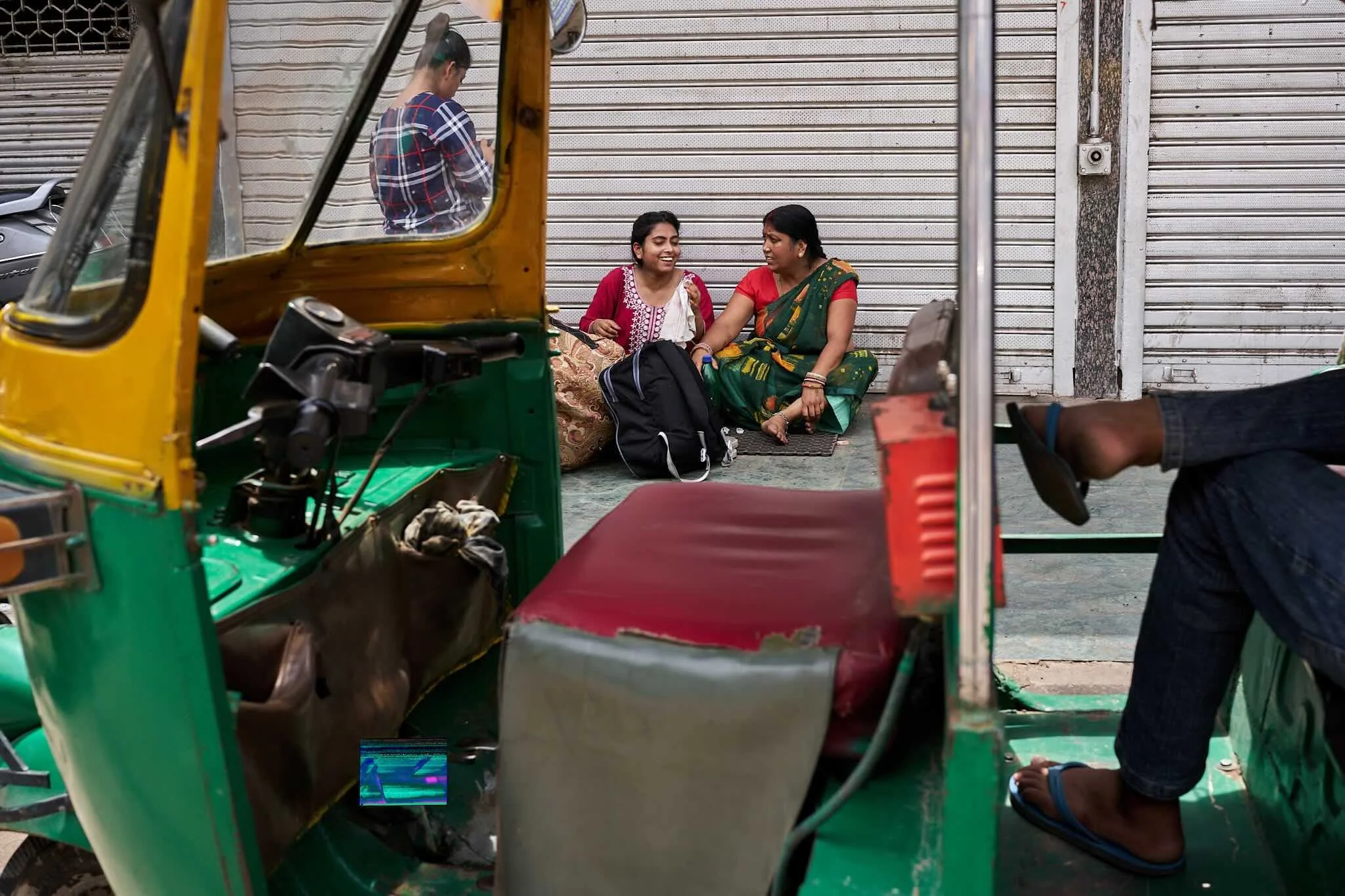 Jerzy Jaworek, Street and Travel Photography, India, Delhi. Two women sitting on the sidewalk in front of a closed metal shutter, smiling and chatting, with an auto rickshaw nearby.
