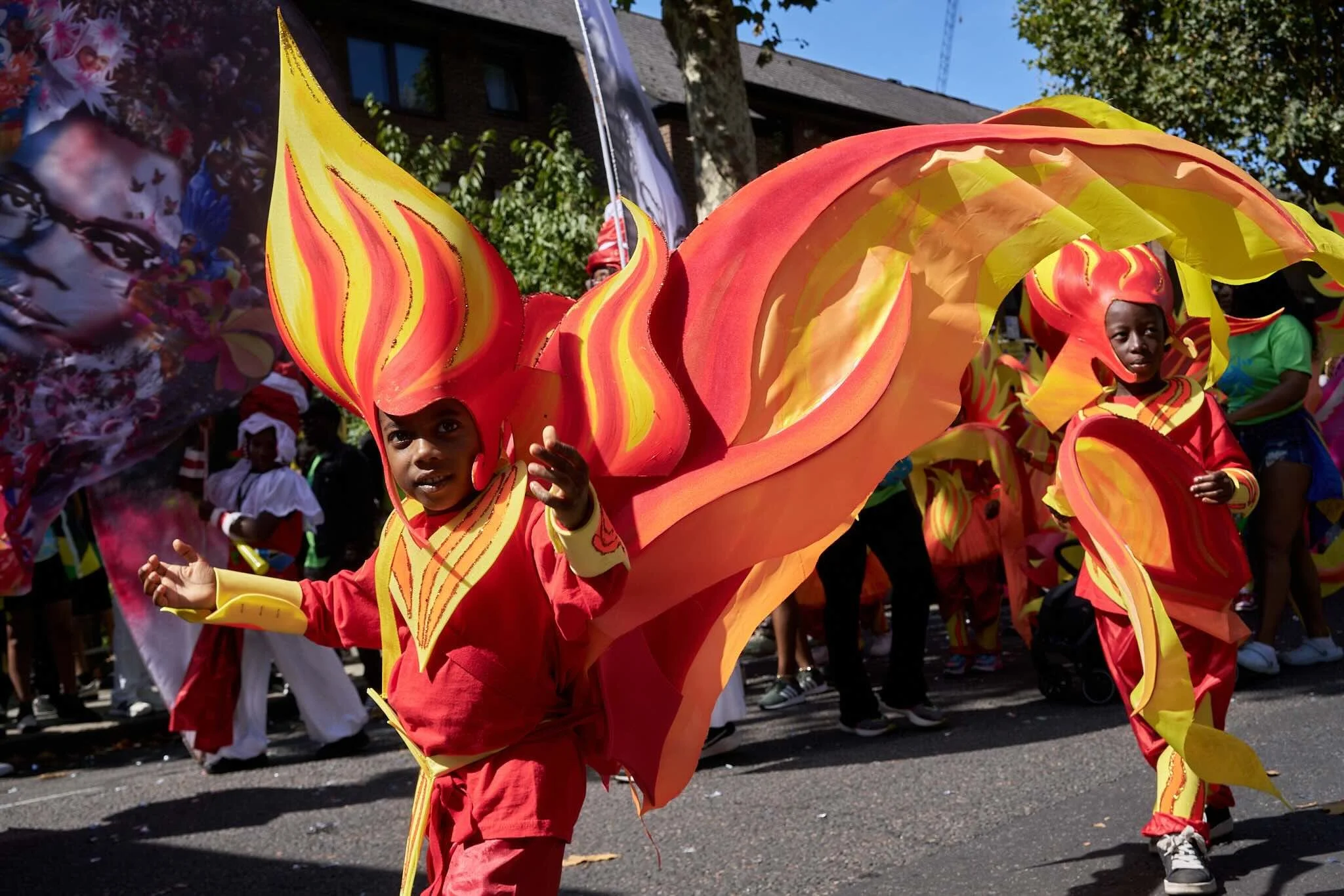 Children dressed in colorful costumes participating in a parade or festival, holding large decorative pieces resembling flames or wings, on a street with spectators and buildings in the background.