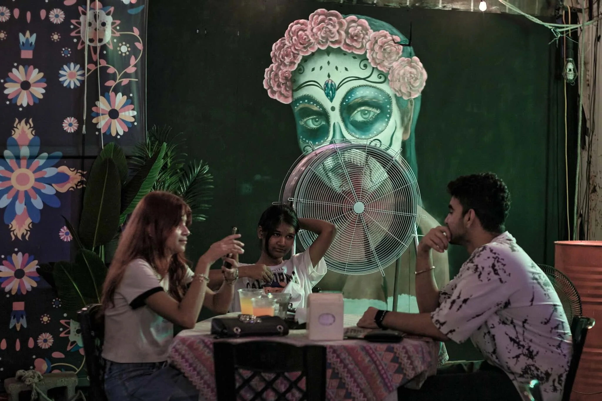 A group of five people sitting at a table in a restaurant or cafe, engaged in conversation. The background features a mural of a face painted with a sugar skull style, surrounded by pink flowers, with tropical plants in front. The setting has a color