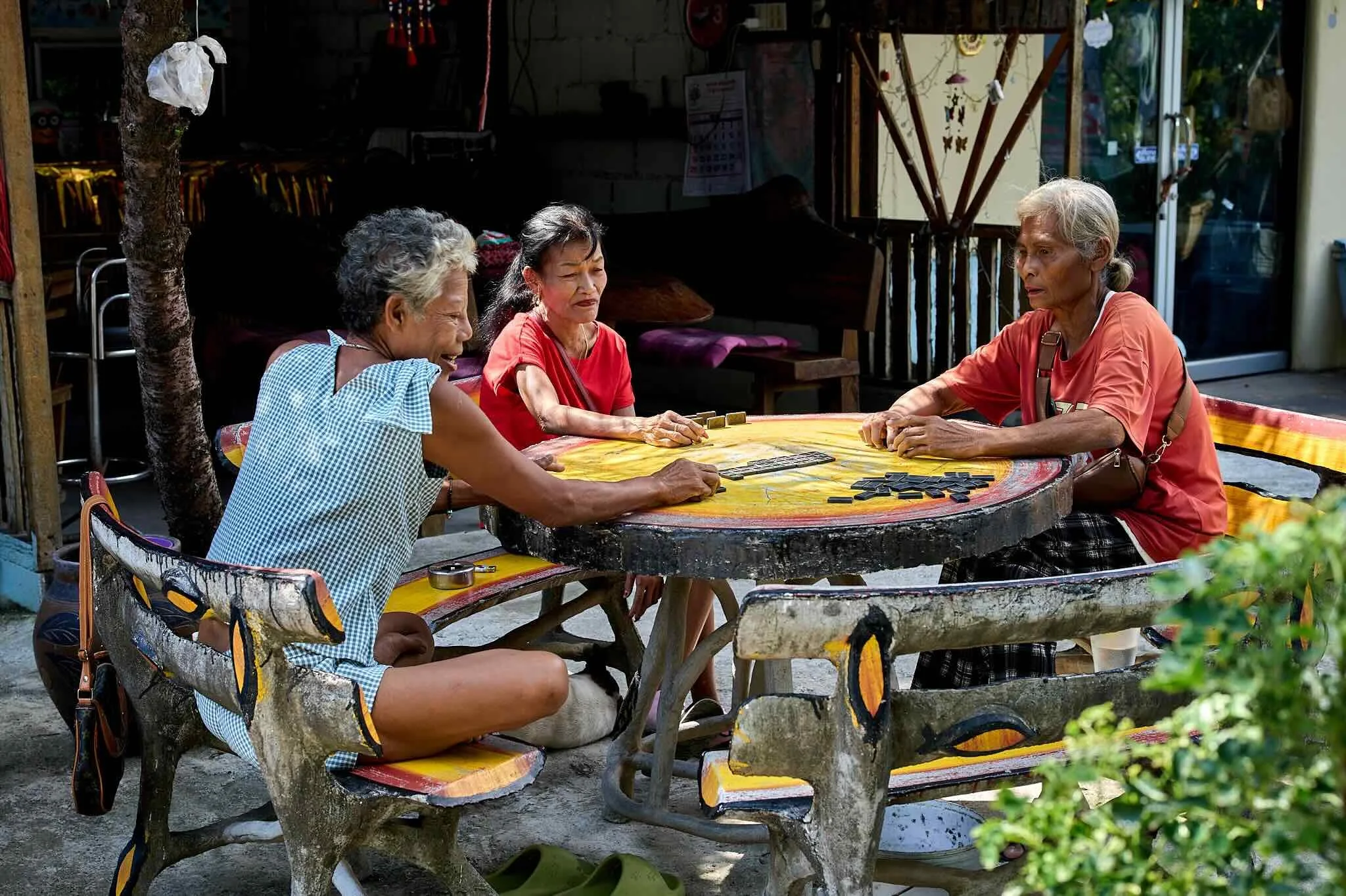 Four senior women sitting around a painted wooden table outside, playing dominoes and engaging in conversation.