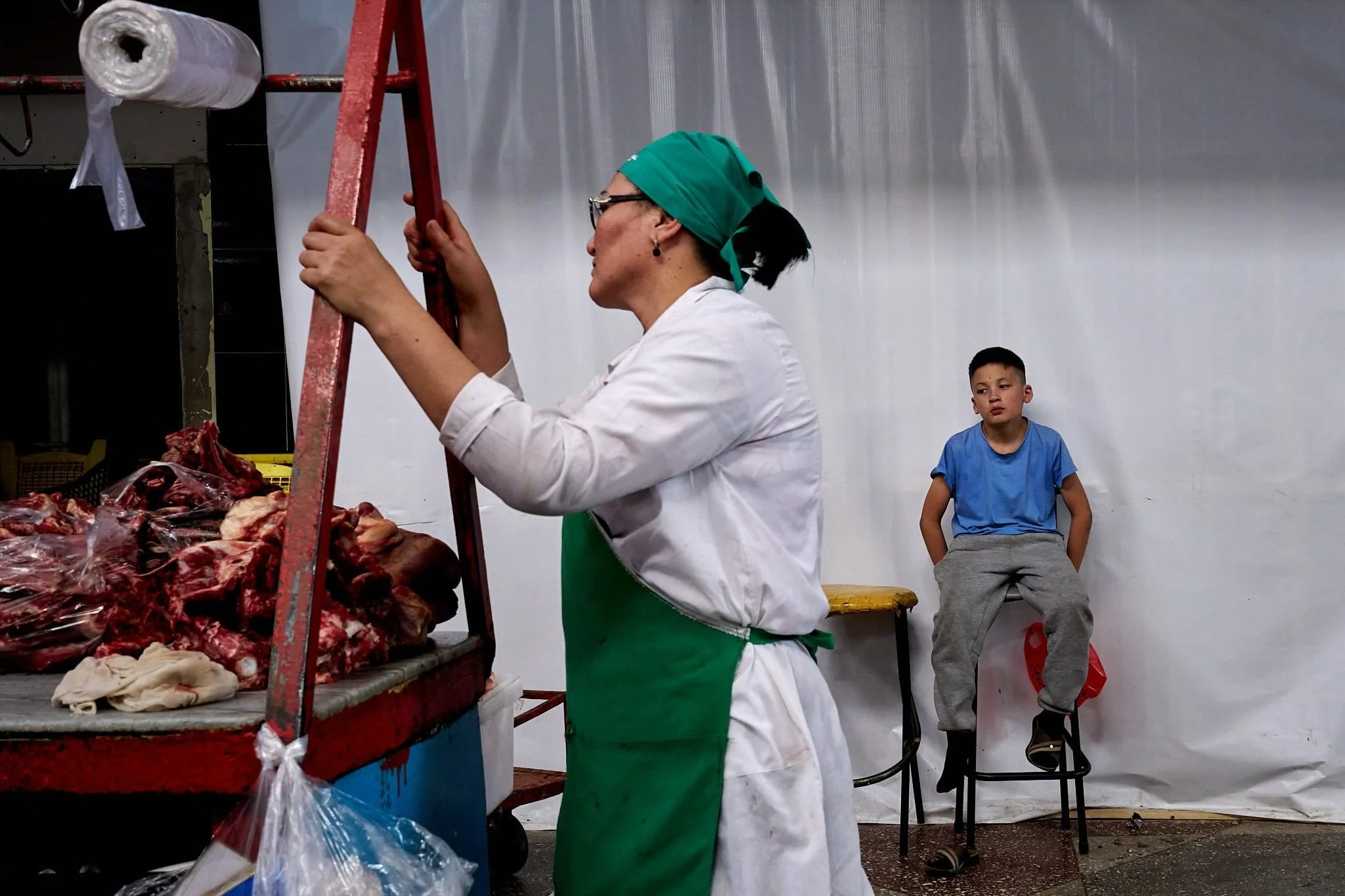 Jerzy Jaworek, Street and Travel Photography, Kazakhstan, Almaty. A woman in green and white apron and green hair covering is inspecting meat at a butcher stand, with a young boy sitting on a stool nearby, against a plain white background.