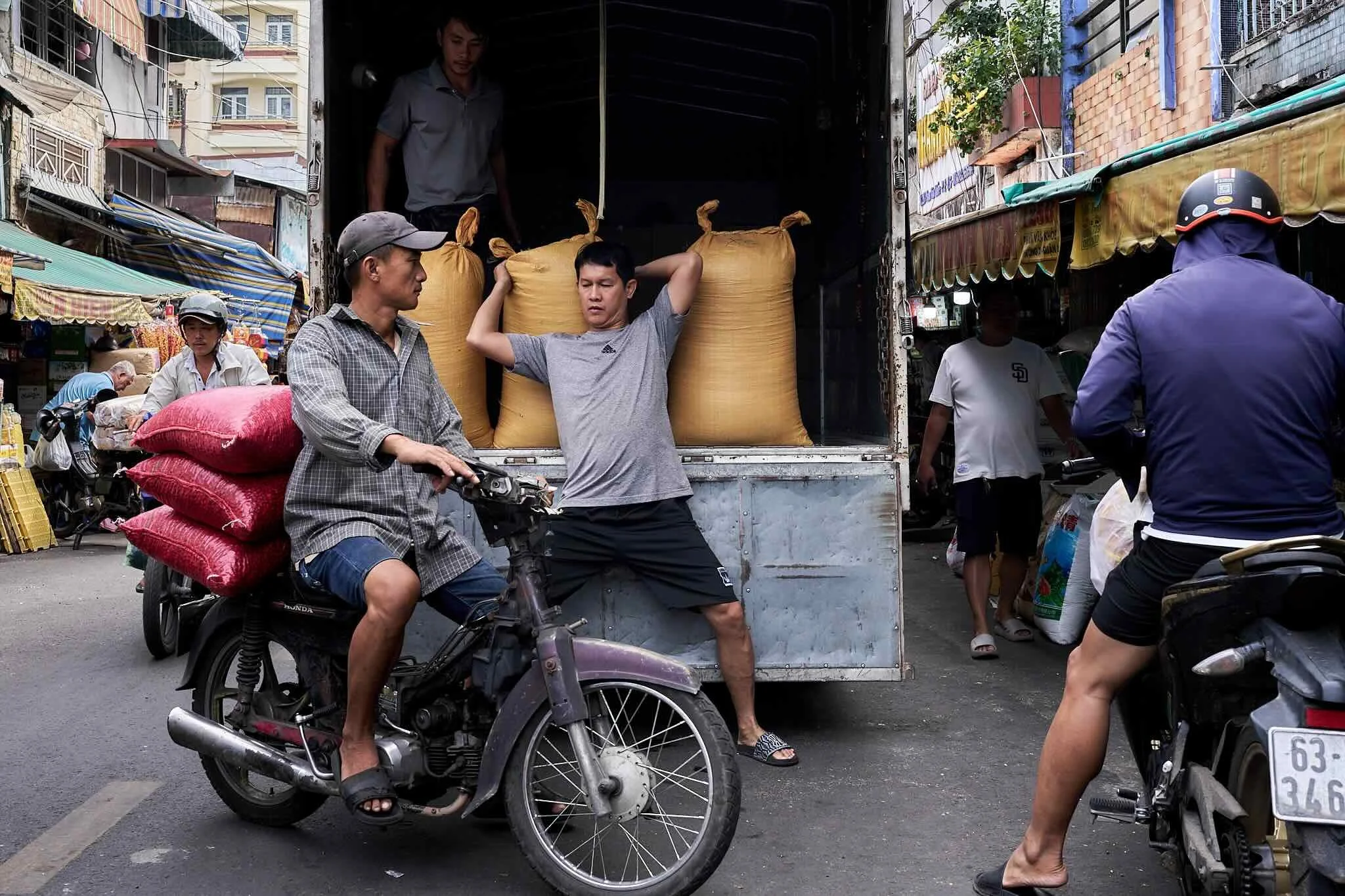 People are loading and unloading goods in a busy outdoor market street, with a man on a motorcycle carrying large red sacks and a truck filled with large brown sacks, surrounded by other individuals and market stalls.