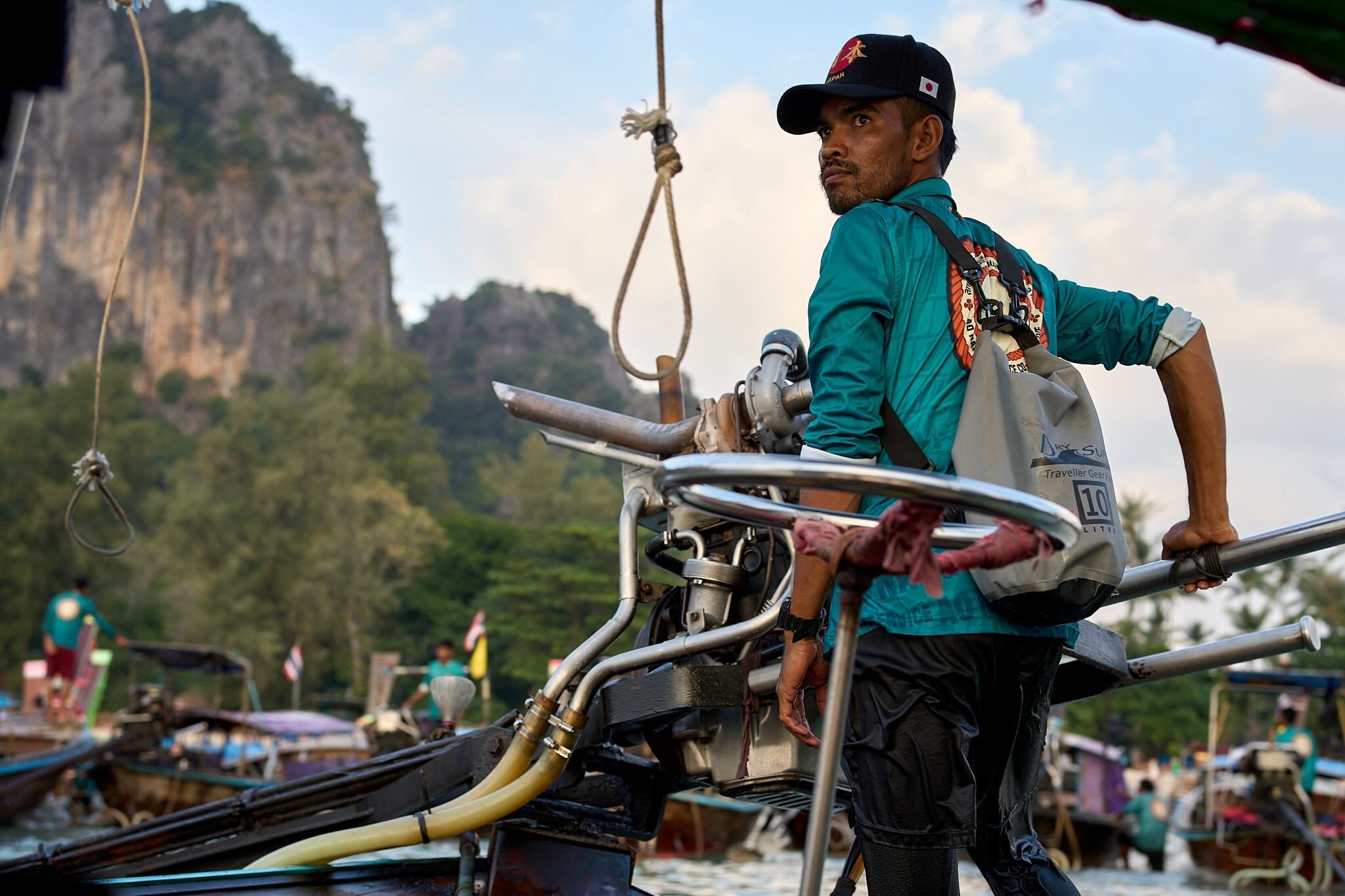 A man wearing a cap and jacket, sitting on a boat, with water and boats in the background.