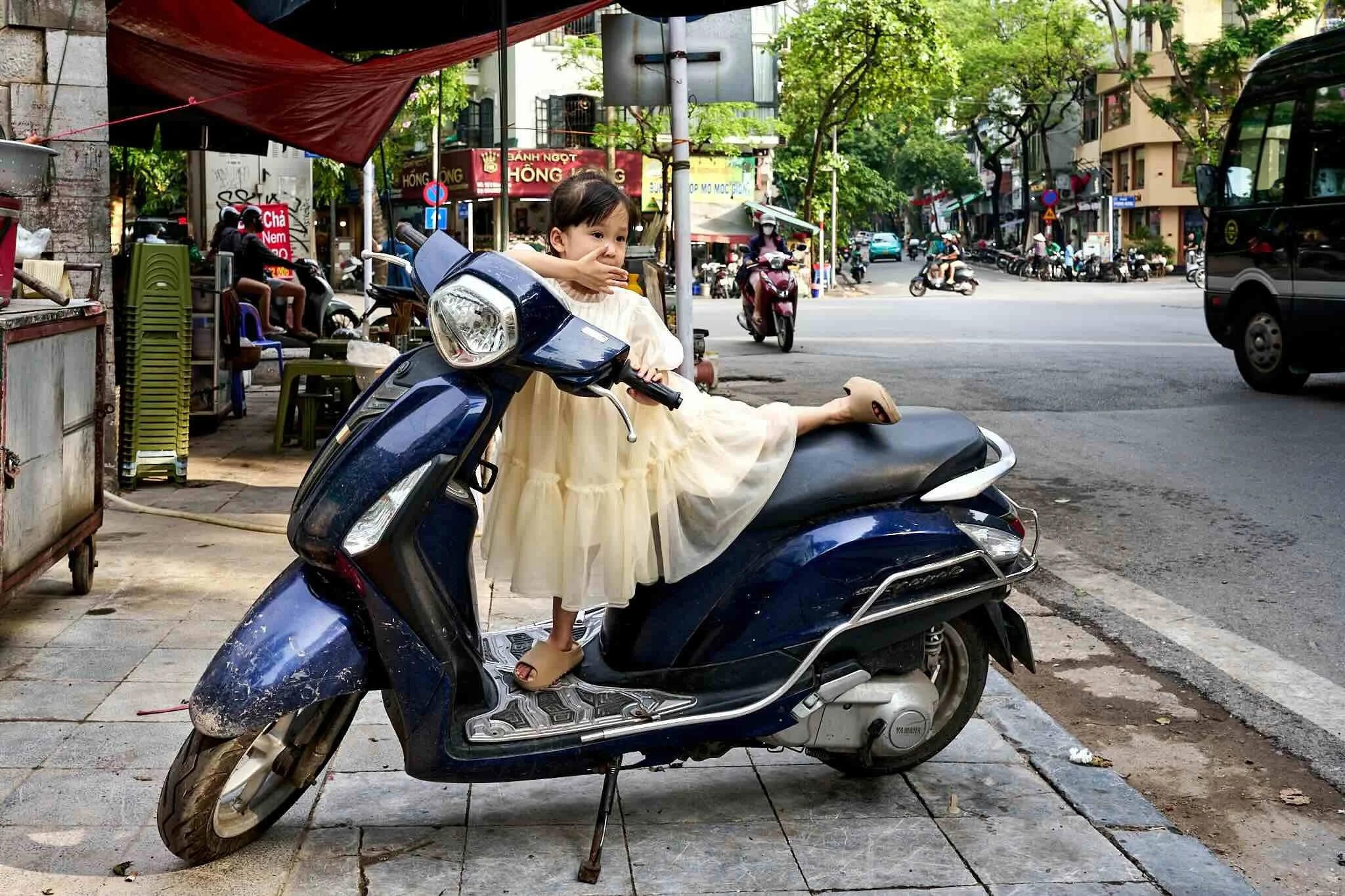 A young girl in a long, light-colored dress and sandals is sitting on a blue scooter parked on a sidewalk in an urban area. She appears to be picking her nose while looking to the side. There are trees, buildings, vehicles, and shops in the backgroun