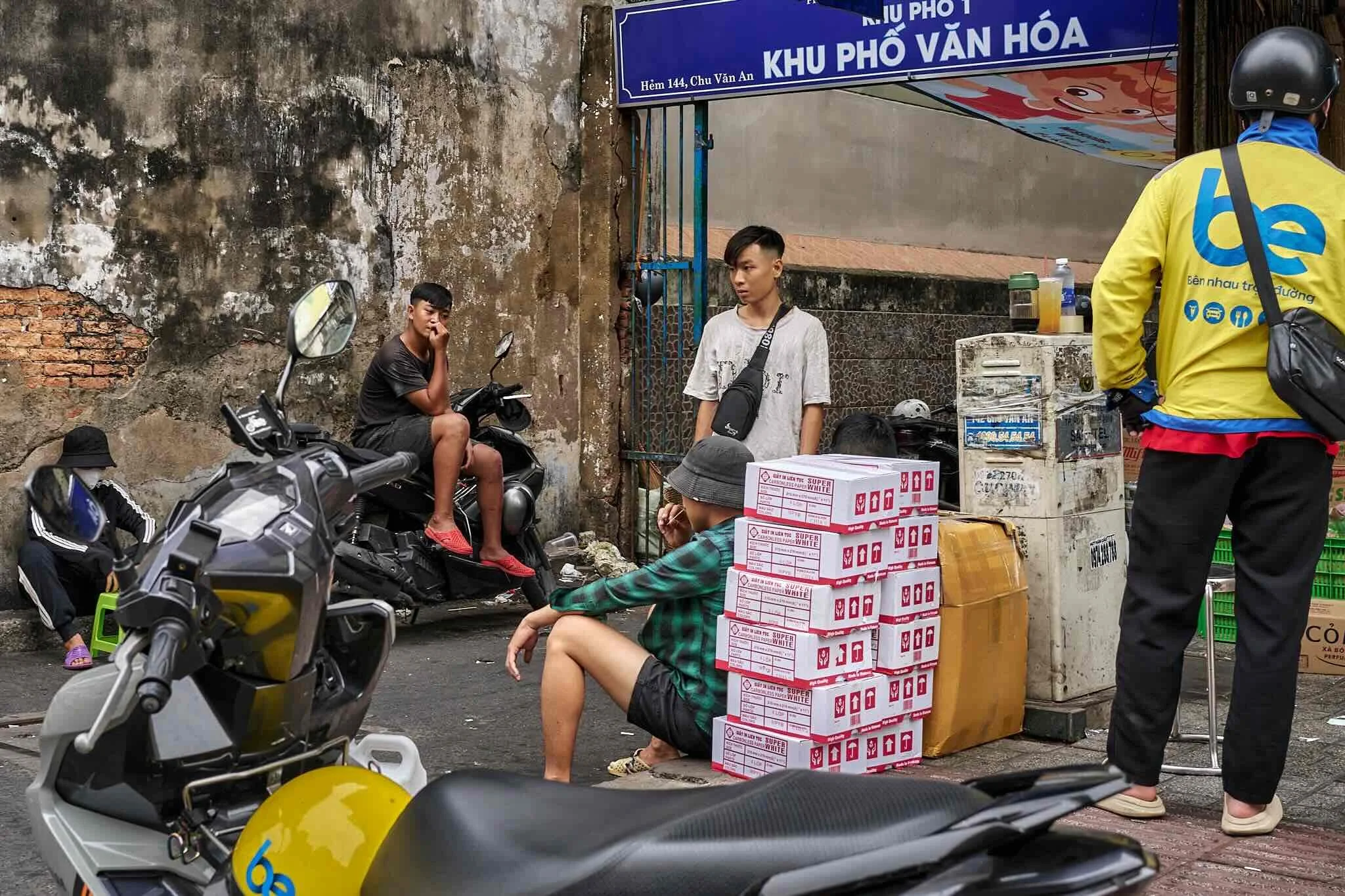 Street scene with young men and motorcycle scooters, boxes stacked near a wall, and a person wearing a yellow jacket riding a bicycle.