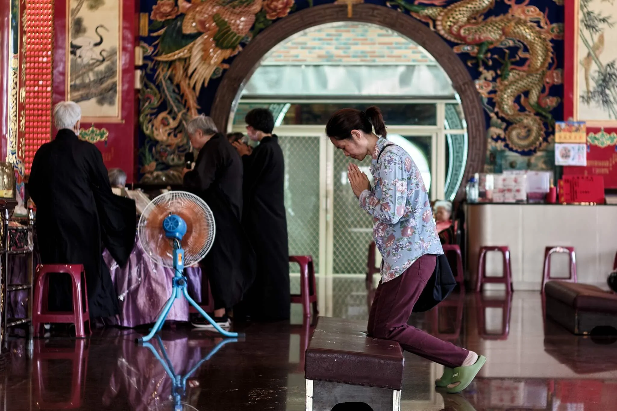 A woman dressed in floral clothing kneeling with folded hands in prayer inside a temple, with people standing in a line behind her.