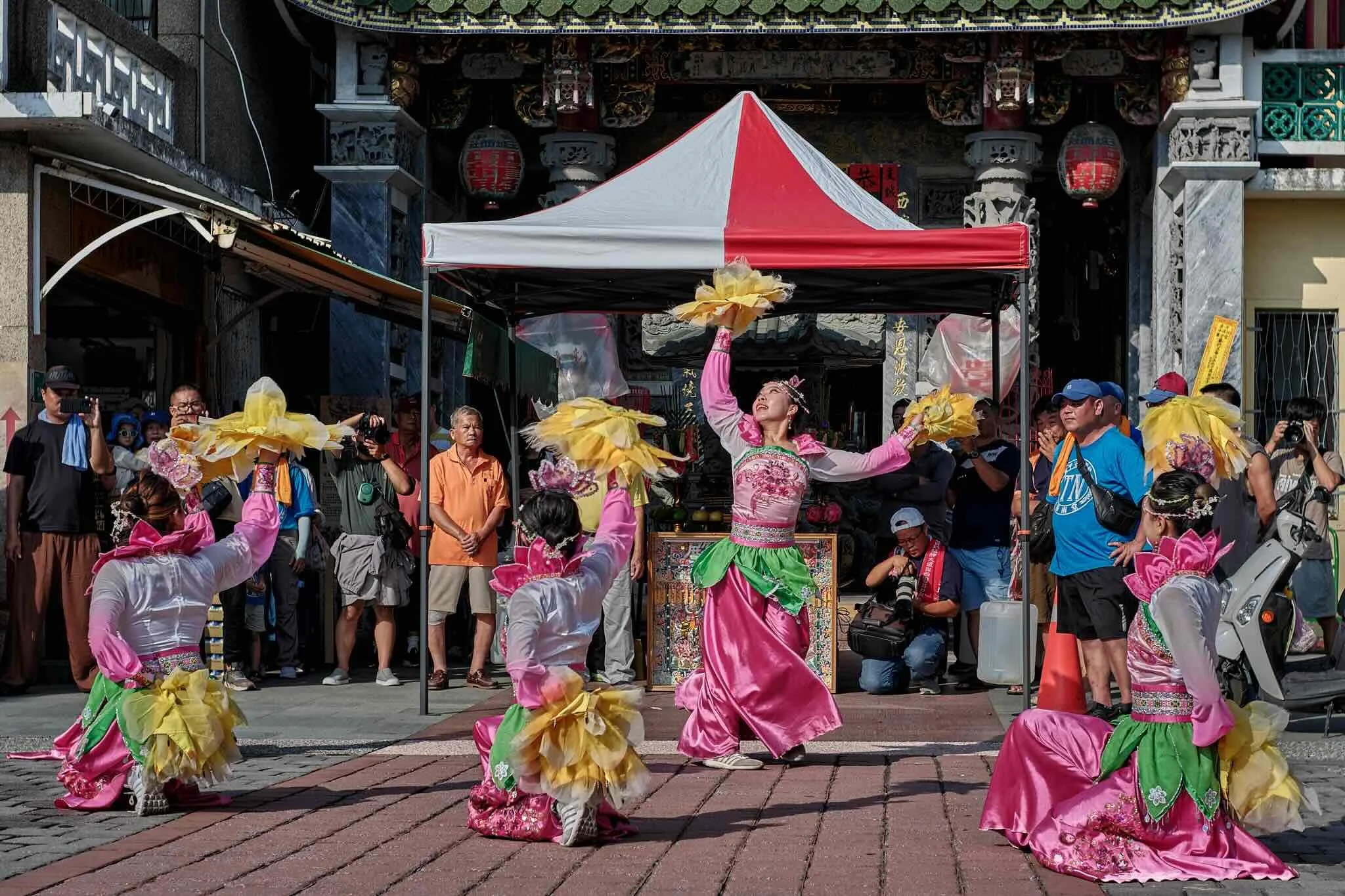 Women in traditional pink and green costumes performing a dance under a red and white canopy, with onlookers gathered around on a sunny day outside a historic building.