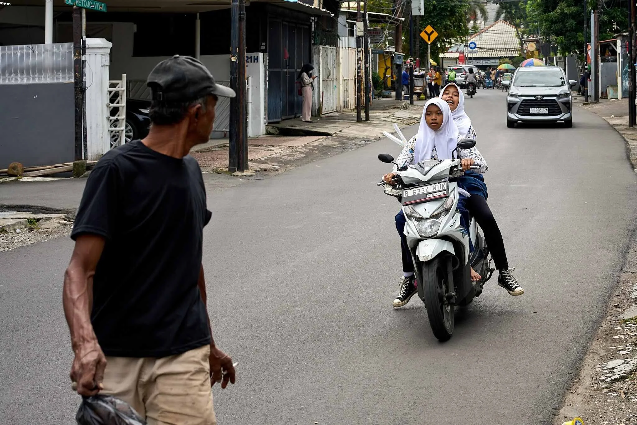 Two young girls wearing white hijabs ride a white scooter on the street, smiling and looking ahead. An older man in a black shirt and beige pants walks towards them. The background shows a lively neighborhood with shops, parked cars, pedestrians, and