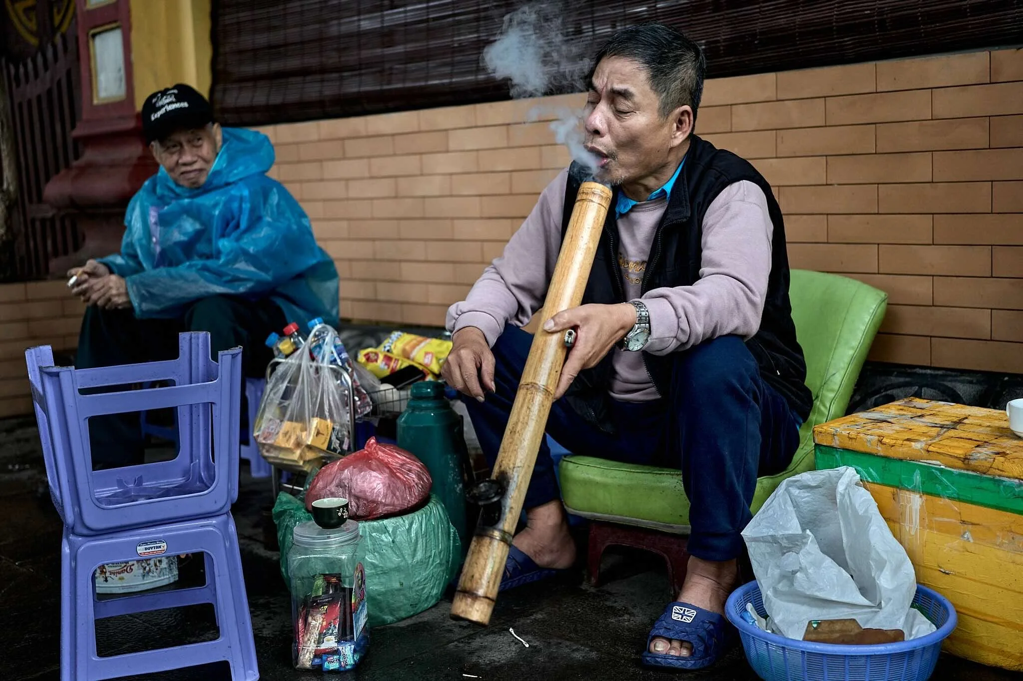 An elderly man with dark hair and mustache playing a bamboo flute, sitting on a green cushion. Another elderly person, wearing a blue raincoat and black cap, is sitting nearby. The scene appears to be outdoors with various plastic bags and containers