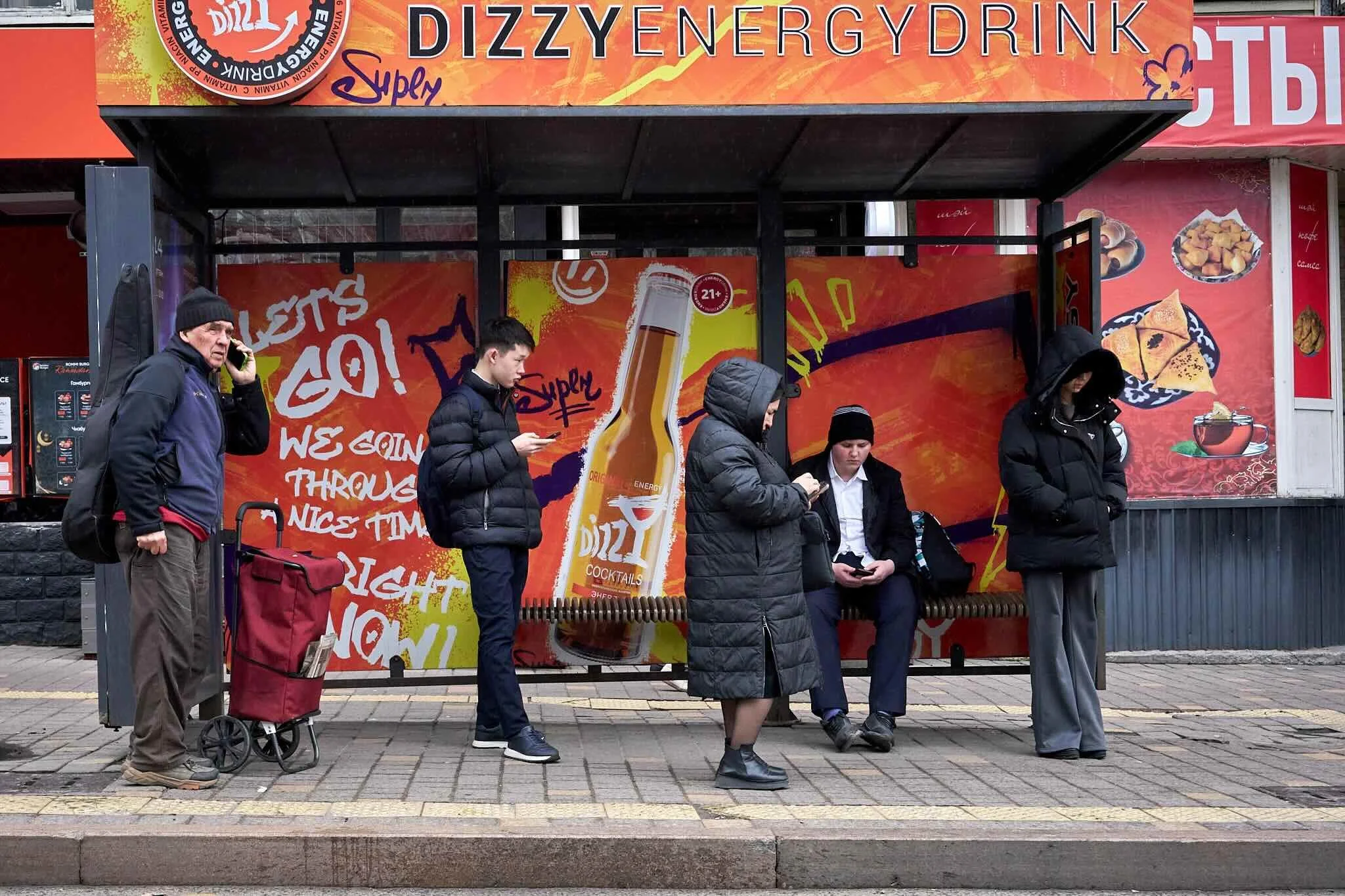 Jerzy Jaworek, Street and Travel Photography, Kazakhstan, Almaty. A bus stop with people standing in line, some looking at their phones, against a bright, colorful advertisement for Dizzy Energy Drink featuring a bottle of the beverage and various fo