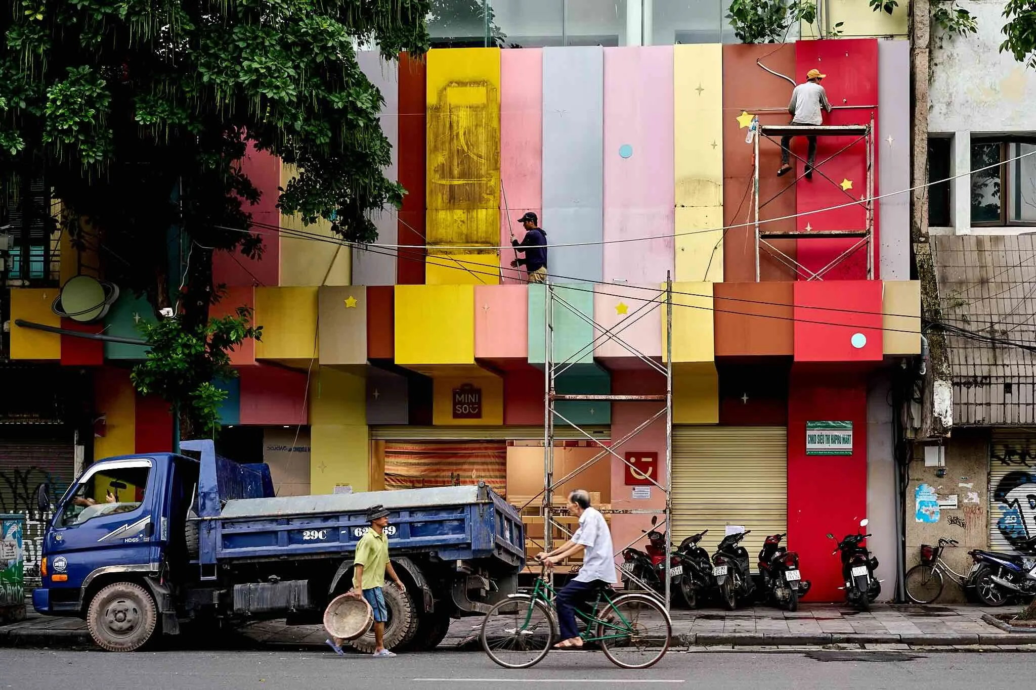 A street scene with a colorful building facade, construction workers renovating the exterior, parked motorbikes, a blue truck, and pedestrians walking on the sidewalk.