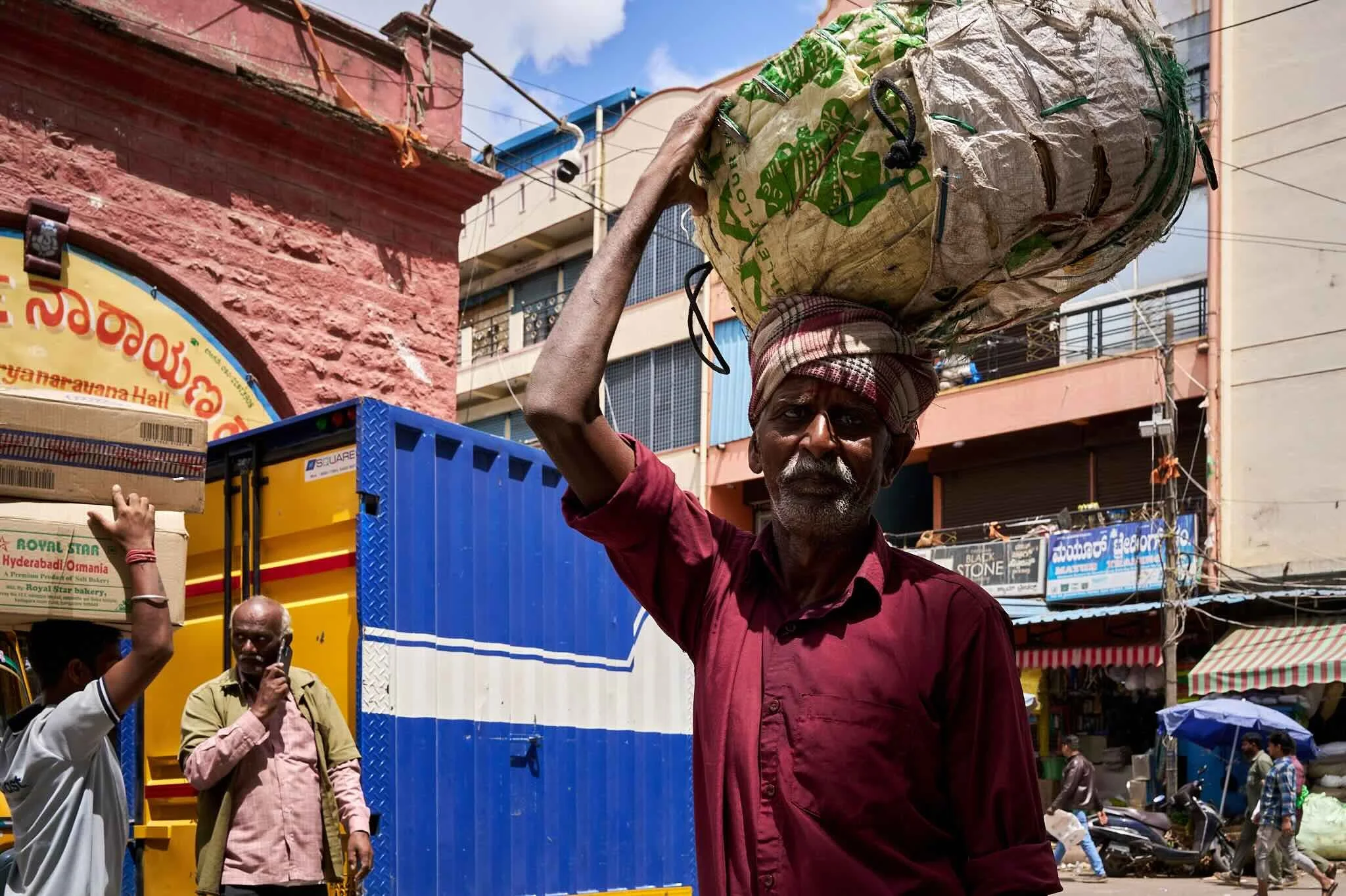 Jerzy Jaworek, Street and Travel Photography, India, Delhi. A man carrying a large bag on his head, walking through a busy street with shops and pedestrians. There are other people visible in the background, with colorful signage and buildings.