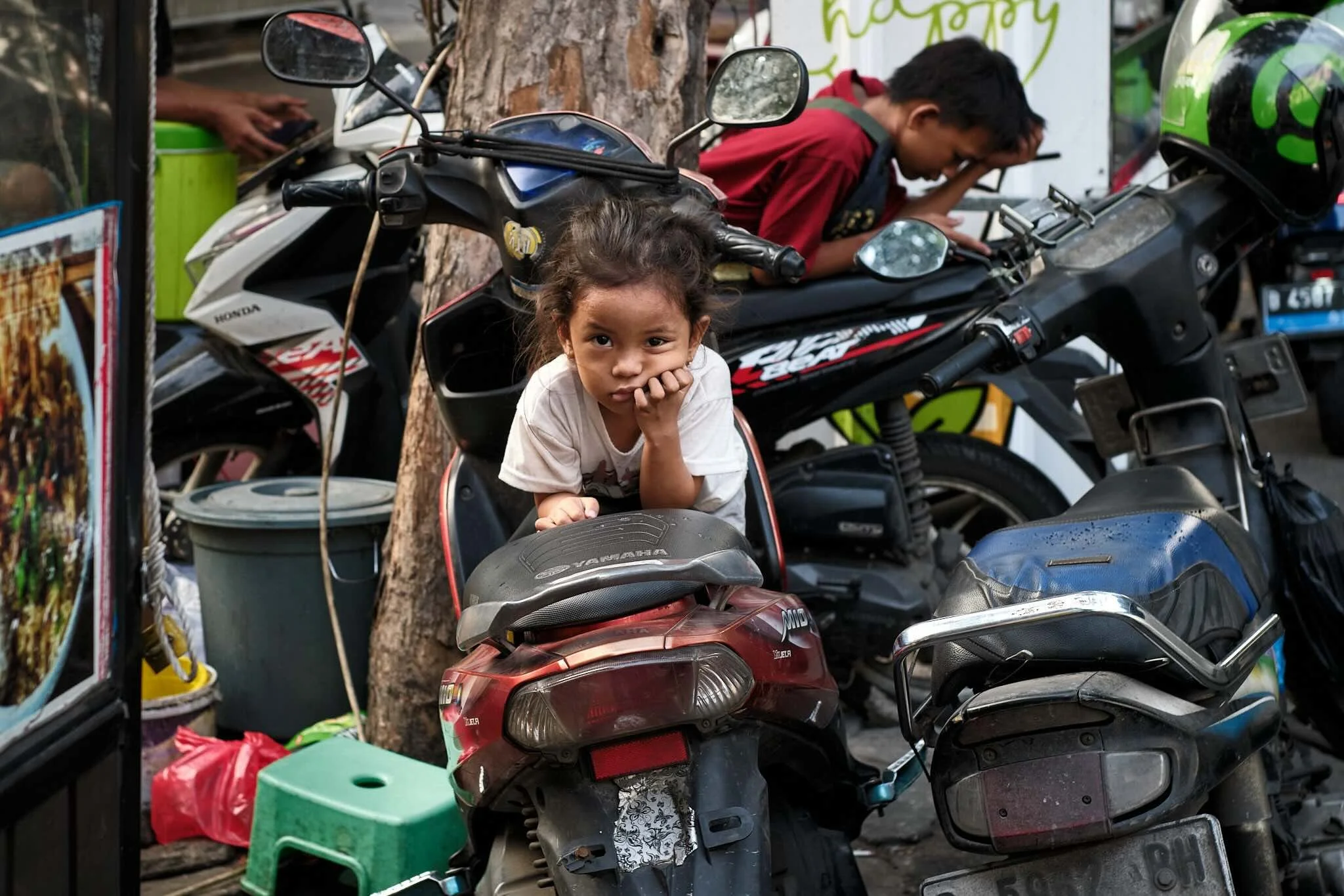 A young girl sitting on a red motorbike with her hand supporting her chin, surrounded by parked motorbikes and scootes, with a young boy leaning over a bike behind her, and colorful posters and items around.