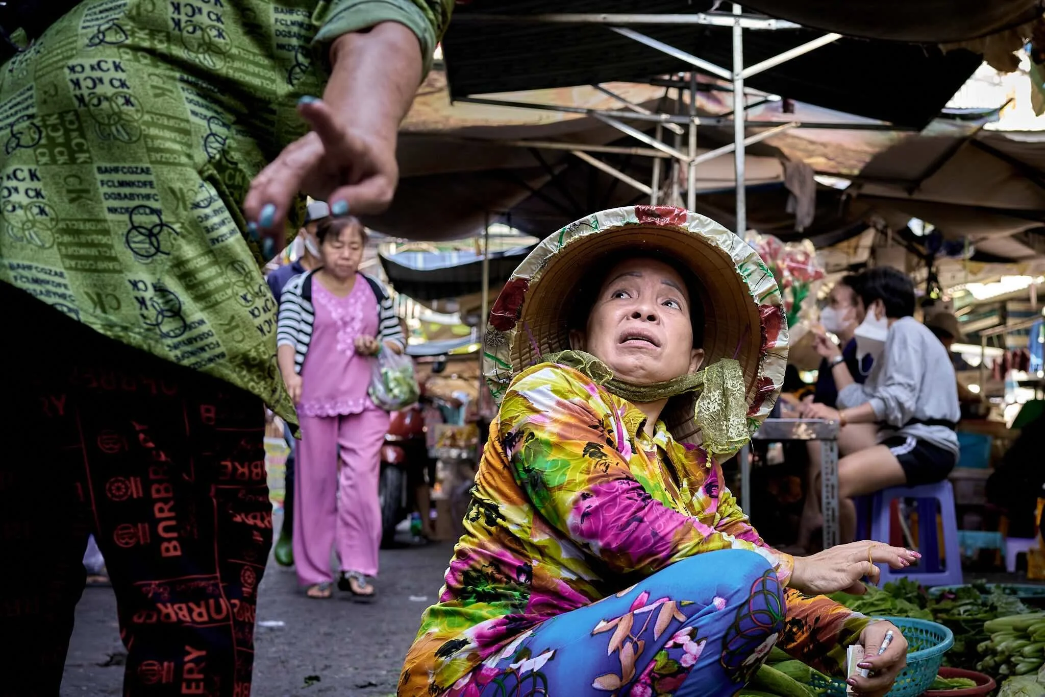 A woman sitting on the ground wearing colorful clothing and a hat, looking up with an expression of concern or surprise at market stalls, with other people shopping in the background under shade tents.