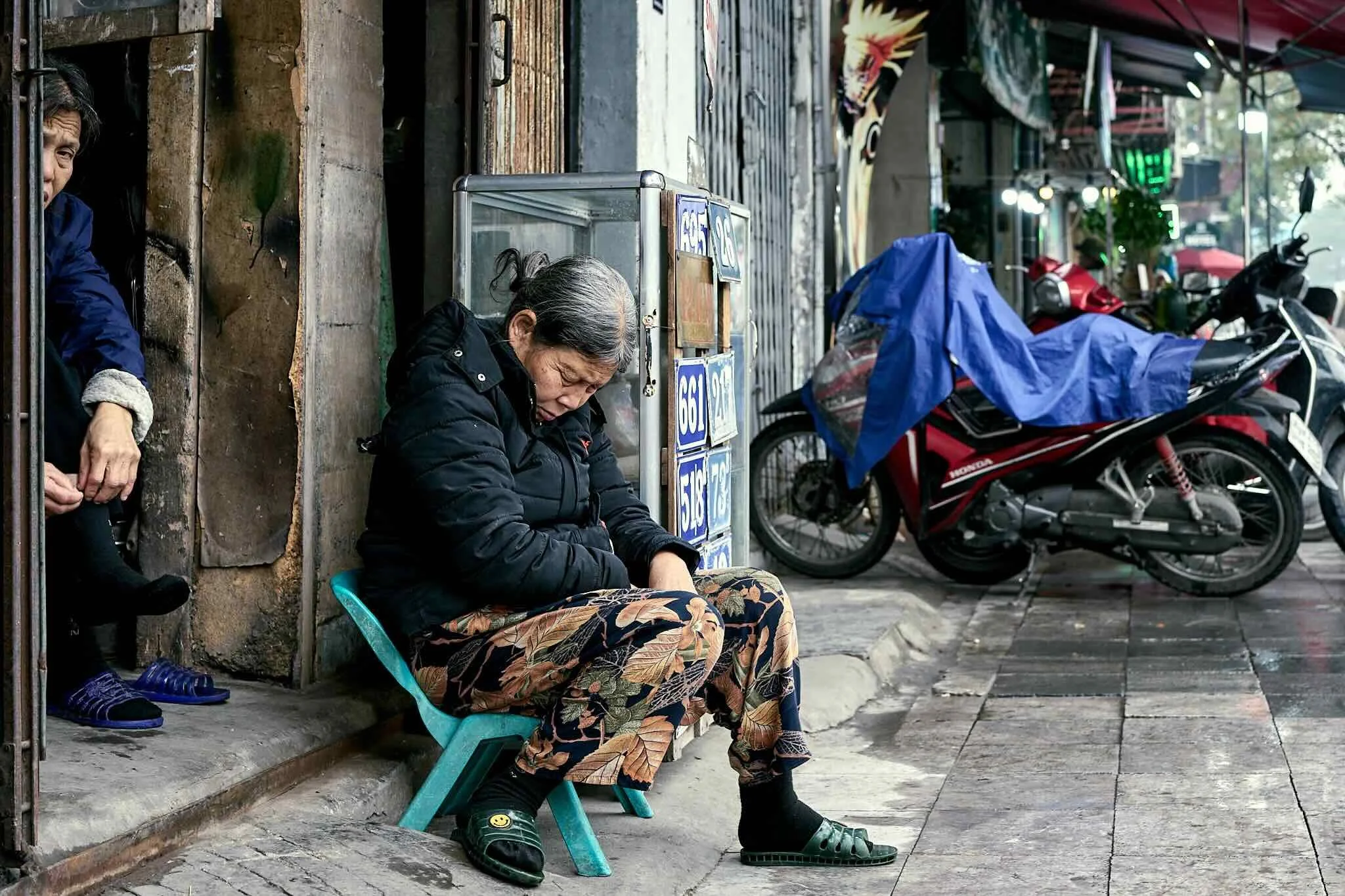 A woman sitting on a small chair on a sidewalk, resting with her eyes closed, dressed in a black jacket and patterned pants, with motorcycles parked nearby and another person partially visible behind her, in an urban street scene.