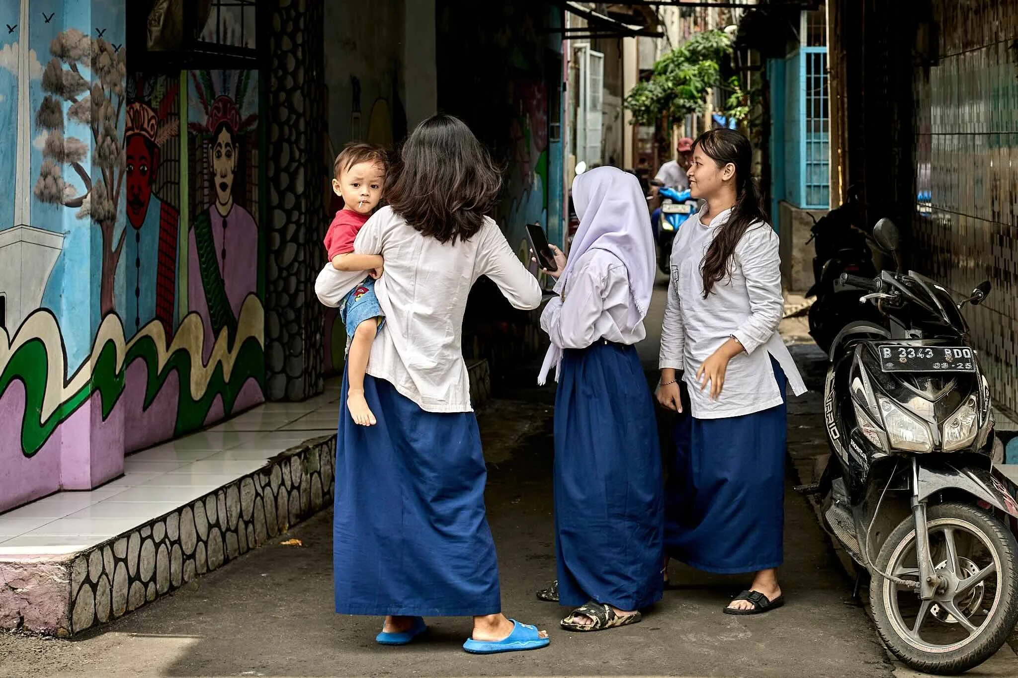 A group of five women and a young boy are standing in an alleyway, chatting. The women are wearing white shirts and long skirts. The boy, held by one woman, is wearing a red shirt and blue shorts and has a lollipop stick in his mouth. The alley has c
