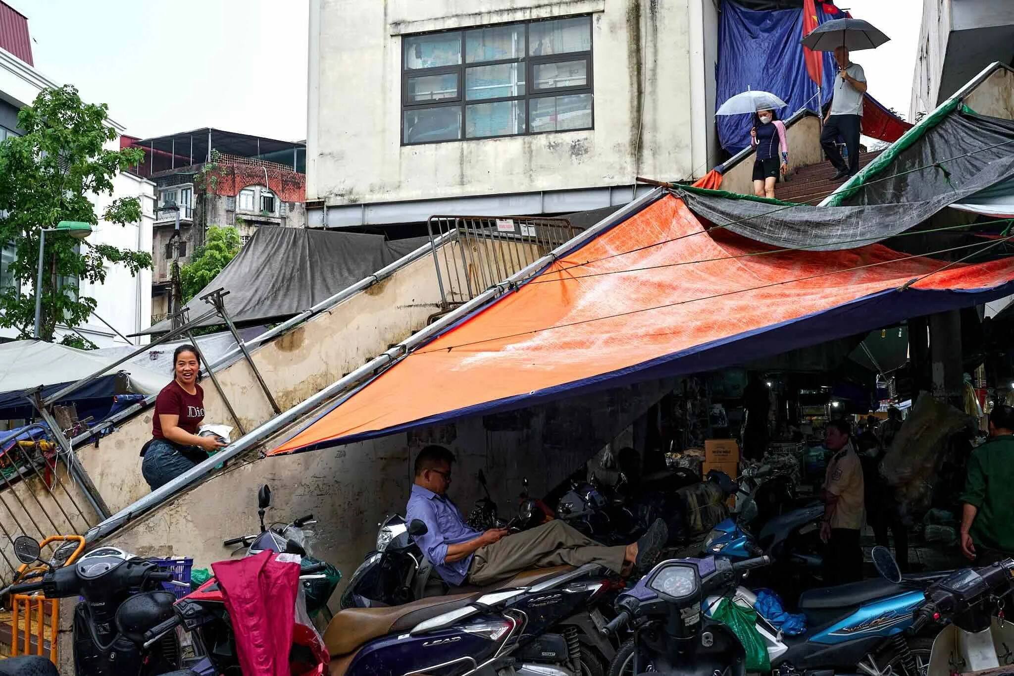 People walking and sitting on motorcycles in a market area with tents and umbrellas, while rain falls on an urban street.