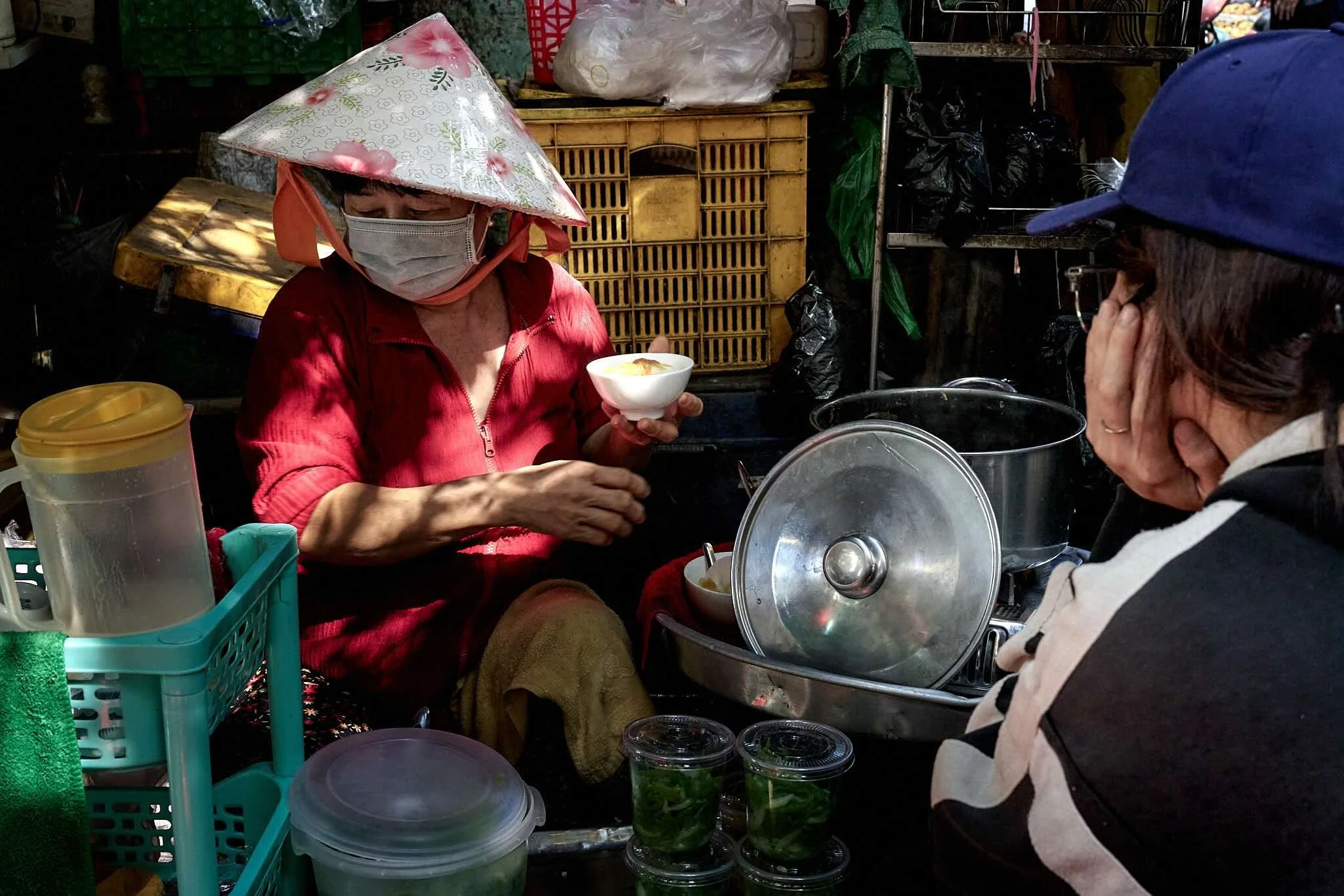 A woman wearing a face mask, a conical hat, and a red jacket is sitting at a street market stall, holding a small bowl of food. Another person wearing a blue cap and glasses is nearby. The stall has various containers, jars of green vegetables, and a