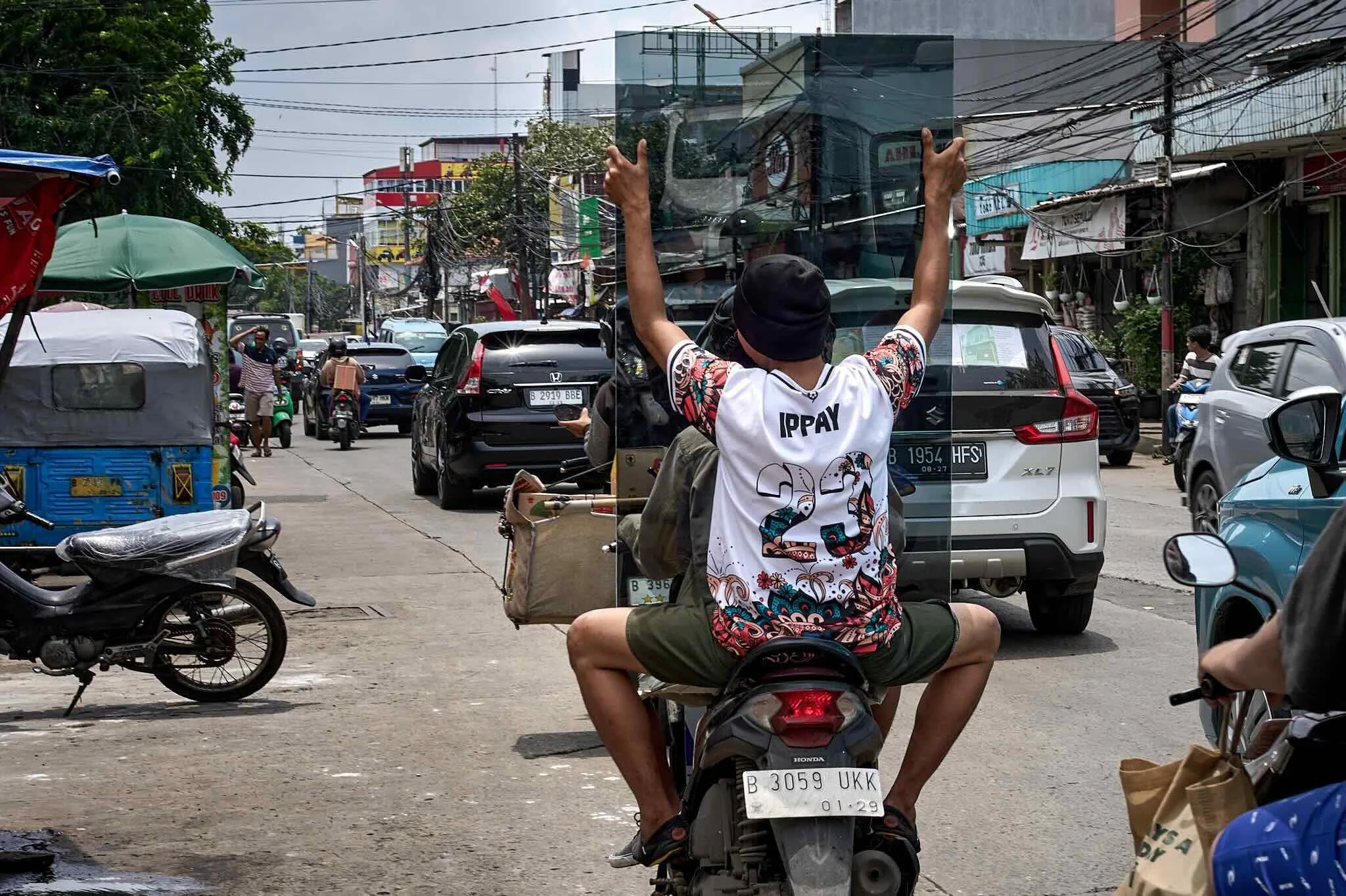 A busy street scene in an urban area with various vehicles, including motorcycles, cars, and a tuk-tuk. A person is sitting on a scooter with a bag, holding up a glass panel, and other pedestrians and motorcyclists are visible in the background. Over