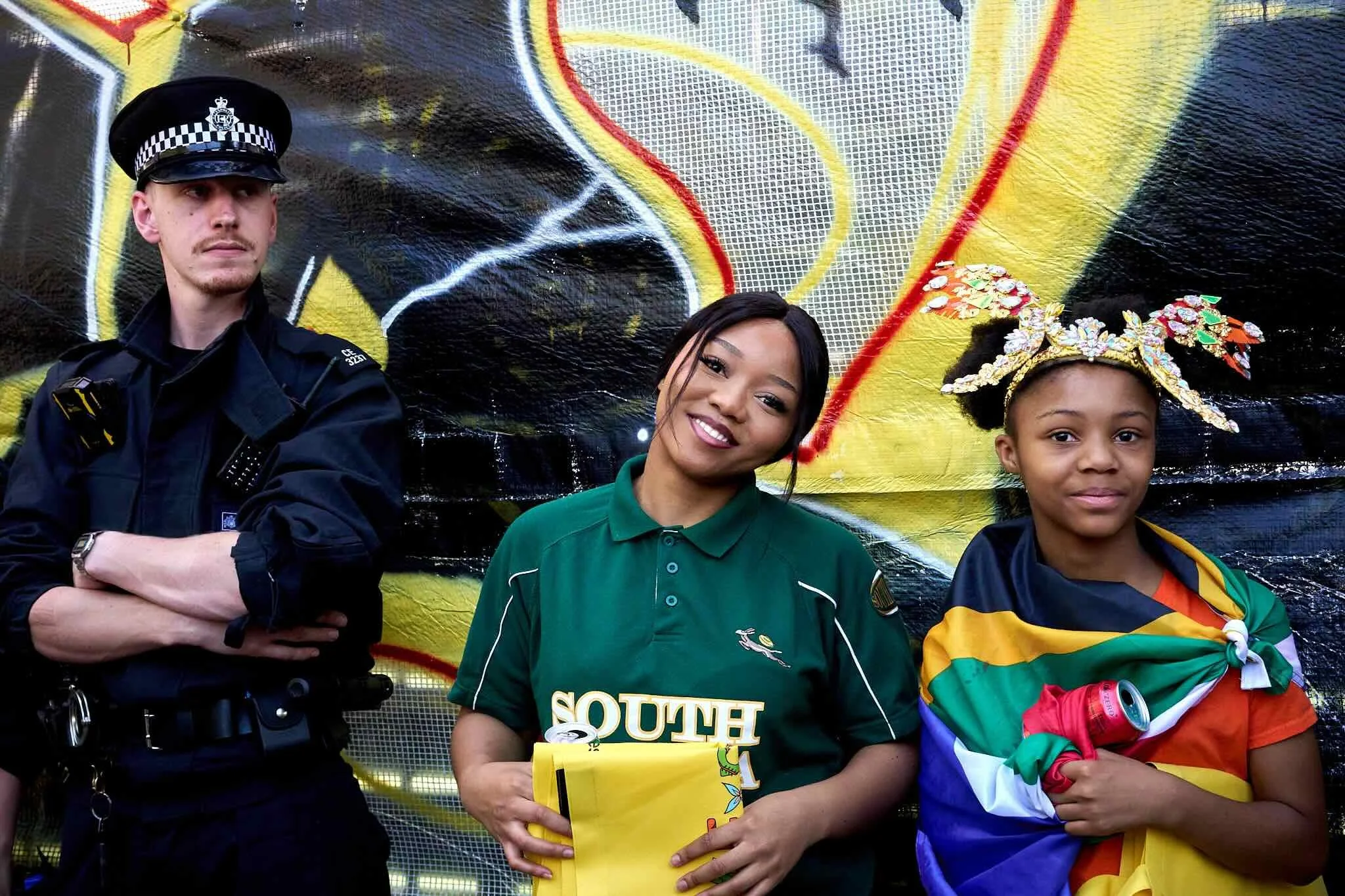 Three people standing in front of a graffiti-covered wall. A police officer on the left, a woman in the middle wearing a green shirt with 'South' written on it, and a young girl on the right wearing a colorful jacket and a decorative headpiece.